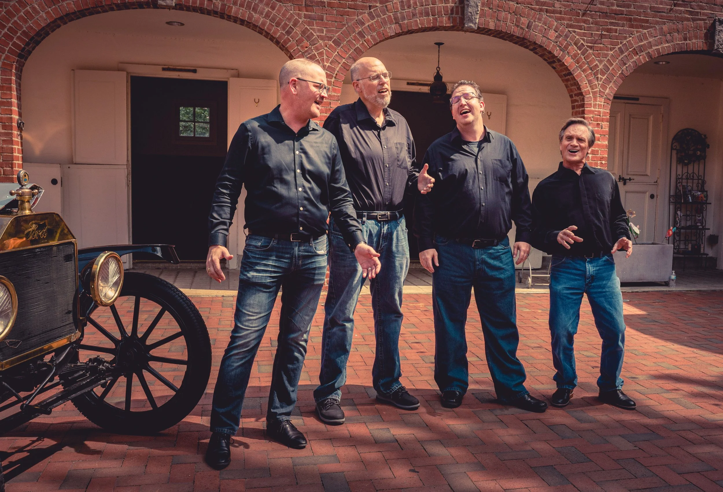 Five men singing together outdoors under brick arches, with vintage car parts partially visible on the left.