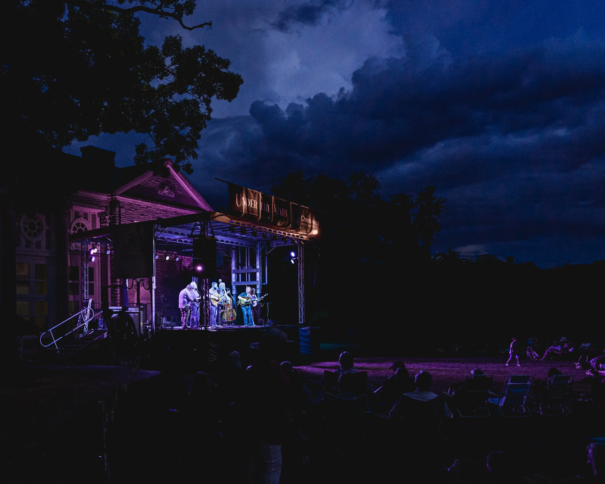 An outdoor concert stage illuminated with purple lights, with a band performing under a dark, cloudy evening sky, surrounded by trees and audience members watching the performance.
