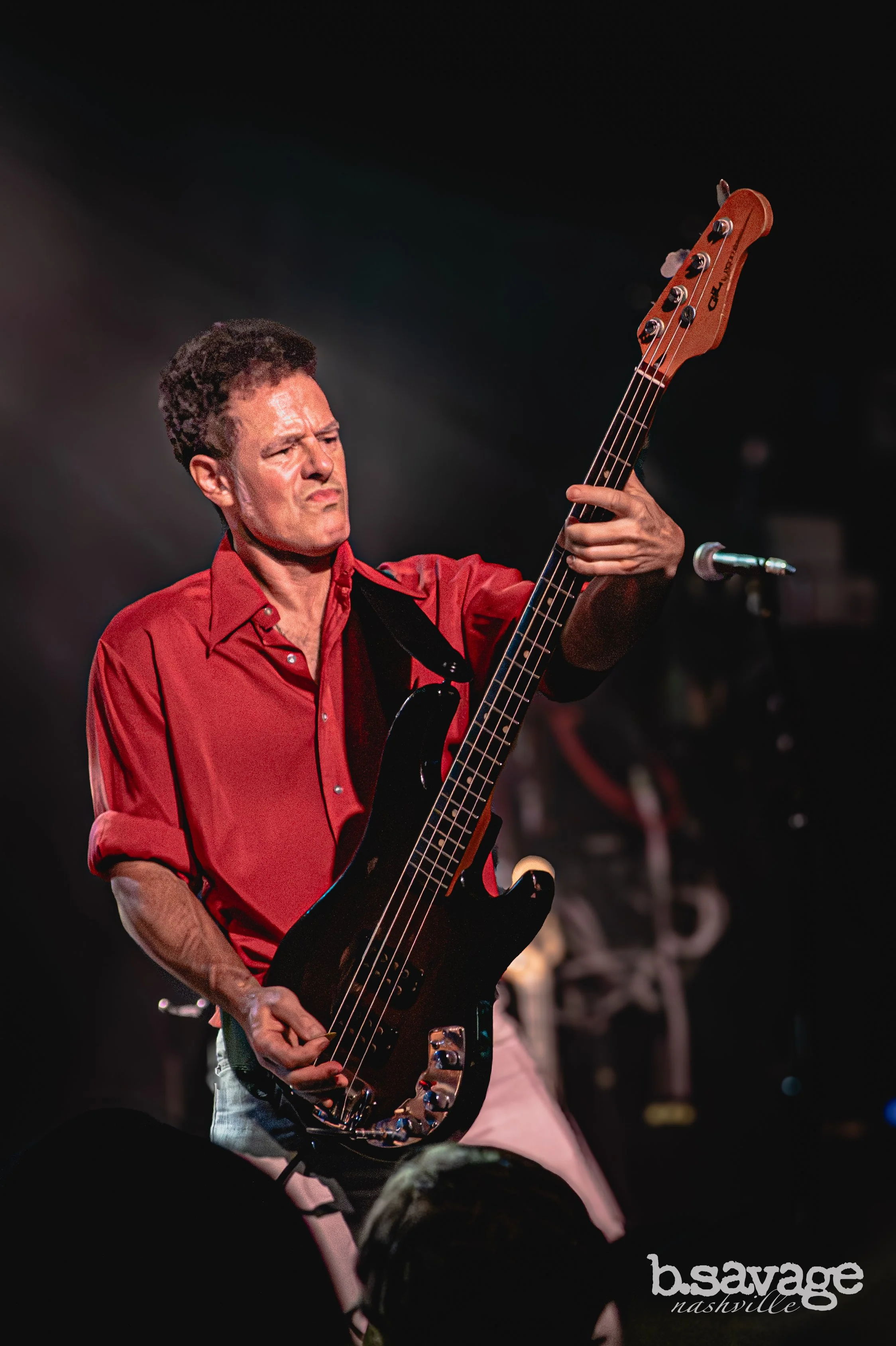 A man in a red shirt playing a bass guitar on stage with a focused expression, in front of an audience at a venue called 'b.savage Nashville'.