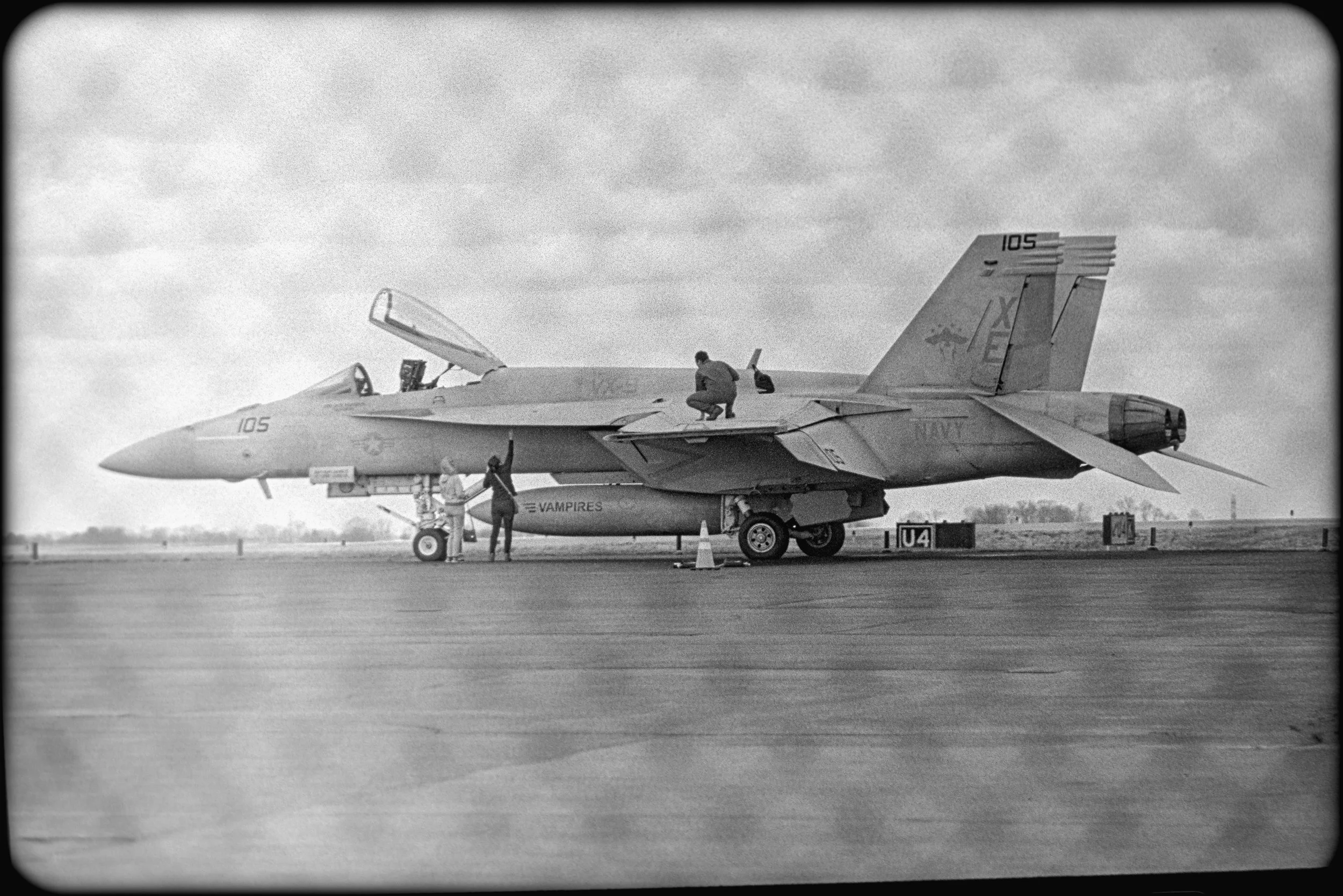 A person kneeling on top of a fighter jet, with four other people standing nearby on an airfield. The jet is marked with the number 105 and has a Navy insignia. The sky is overcast.
