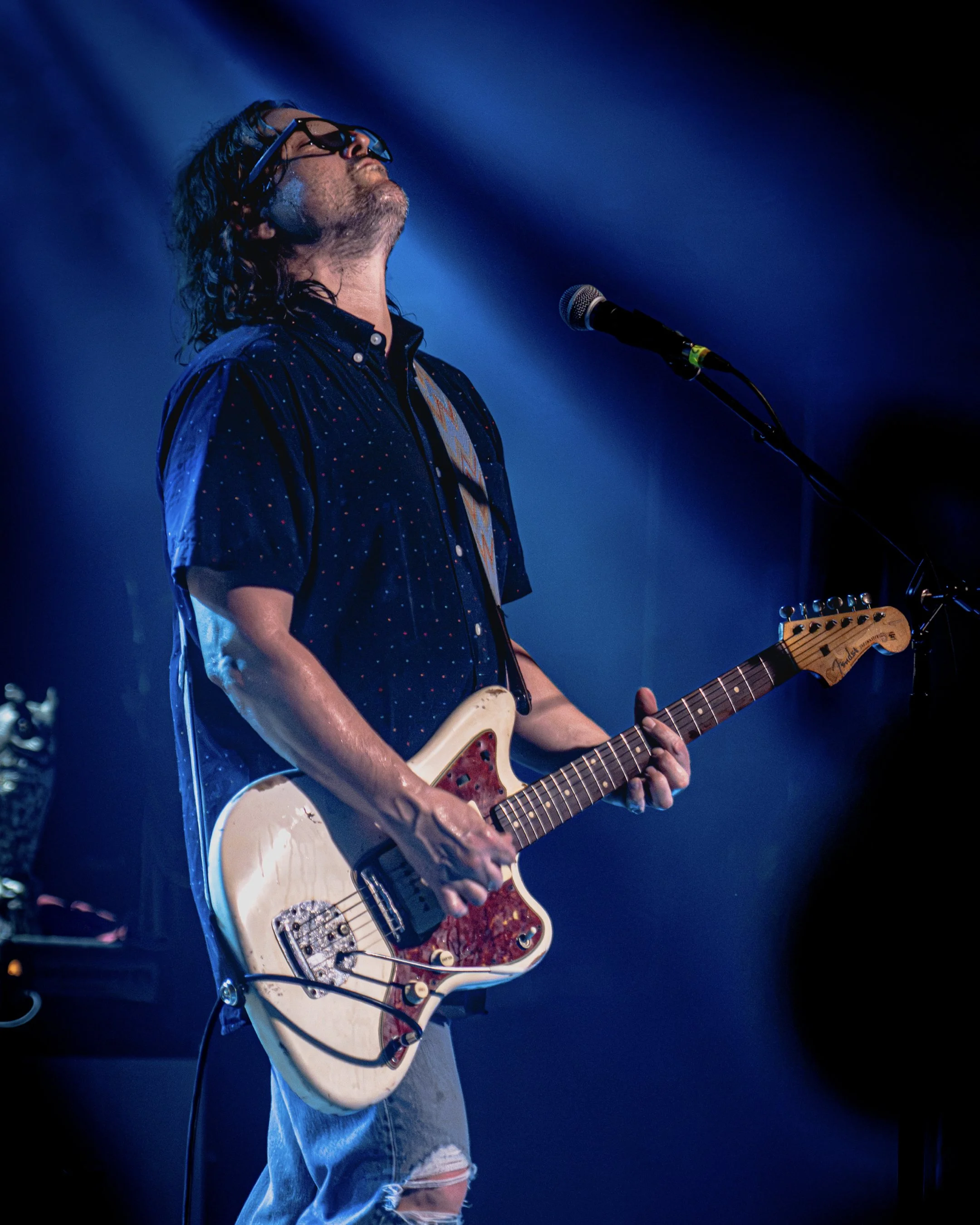 A man with long curly hair singing passionately into a microphone while playing an electric guitar on stage under blue stage lighting.