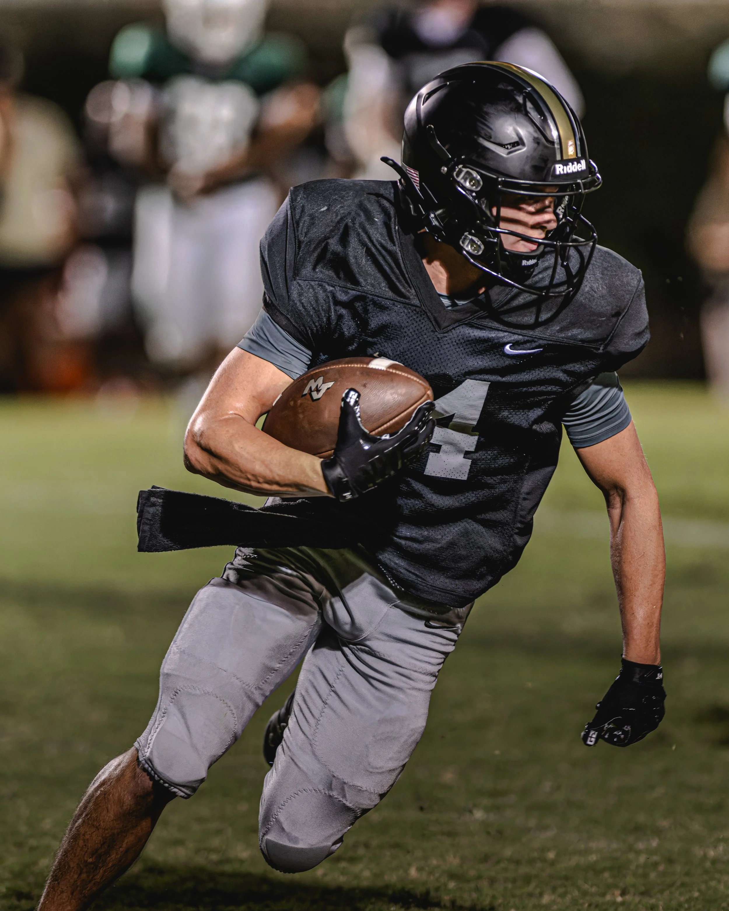 A football player in black helmet and black and gray uniform running with the ball on a football field at night.