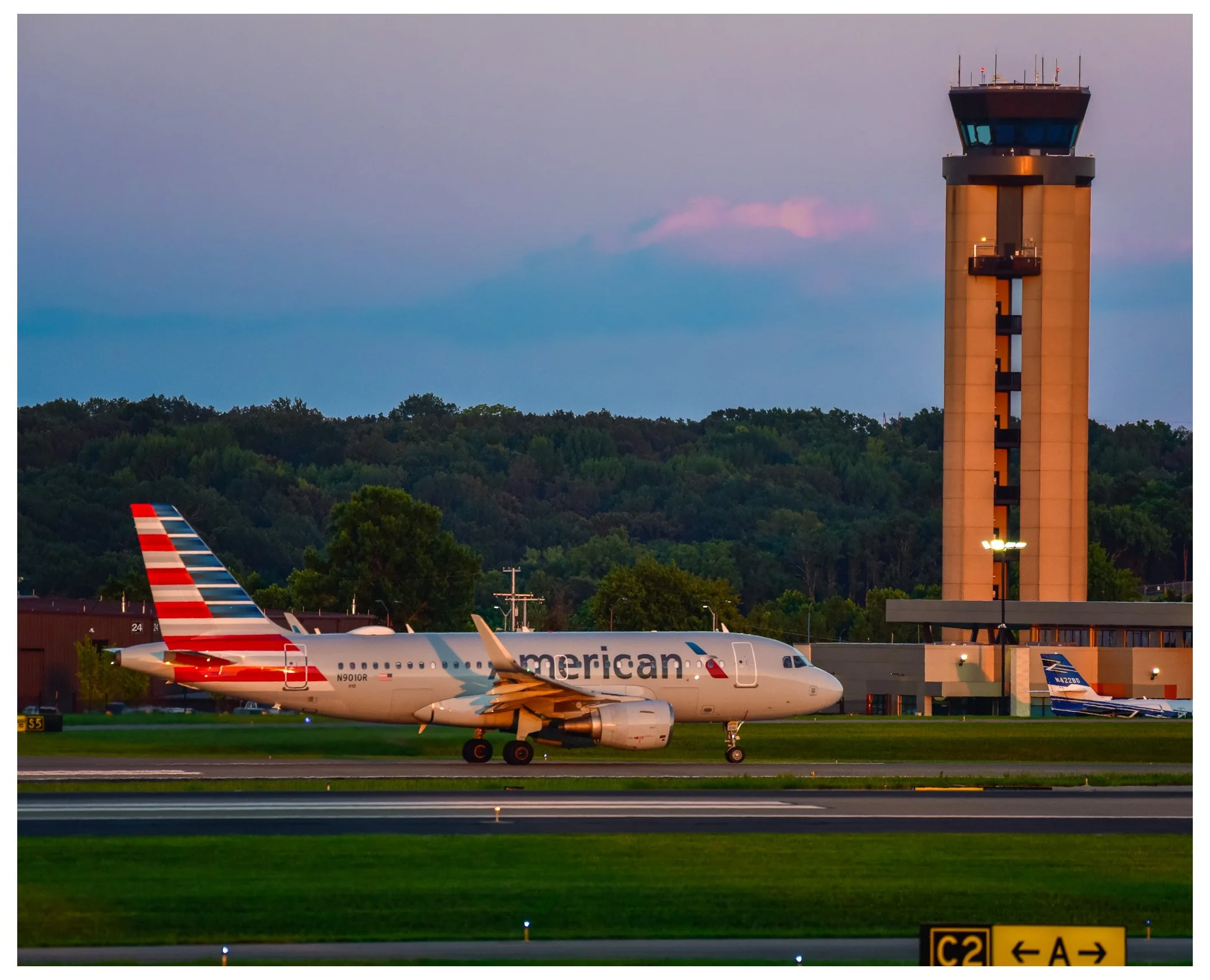 An airport scene at dusk featuring an American Airlines airplane on the runway with the tail fin visible displaying the red, white, and blue stripes. In the background, there is an air traffic control tower and a lush green hillside. The airport runw