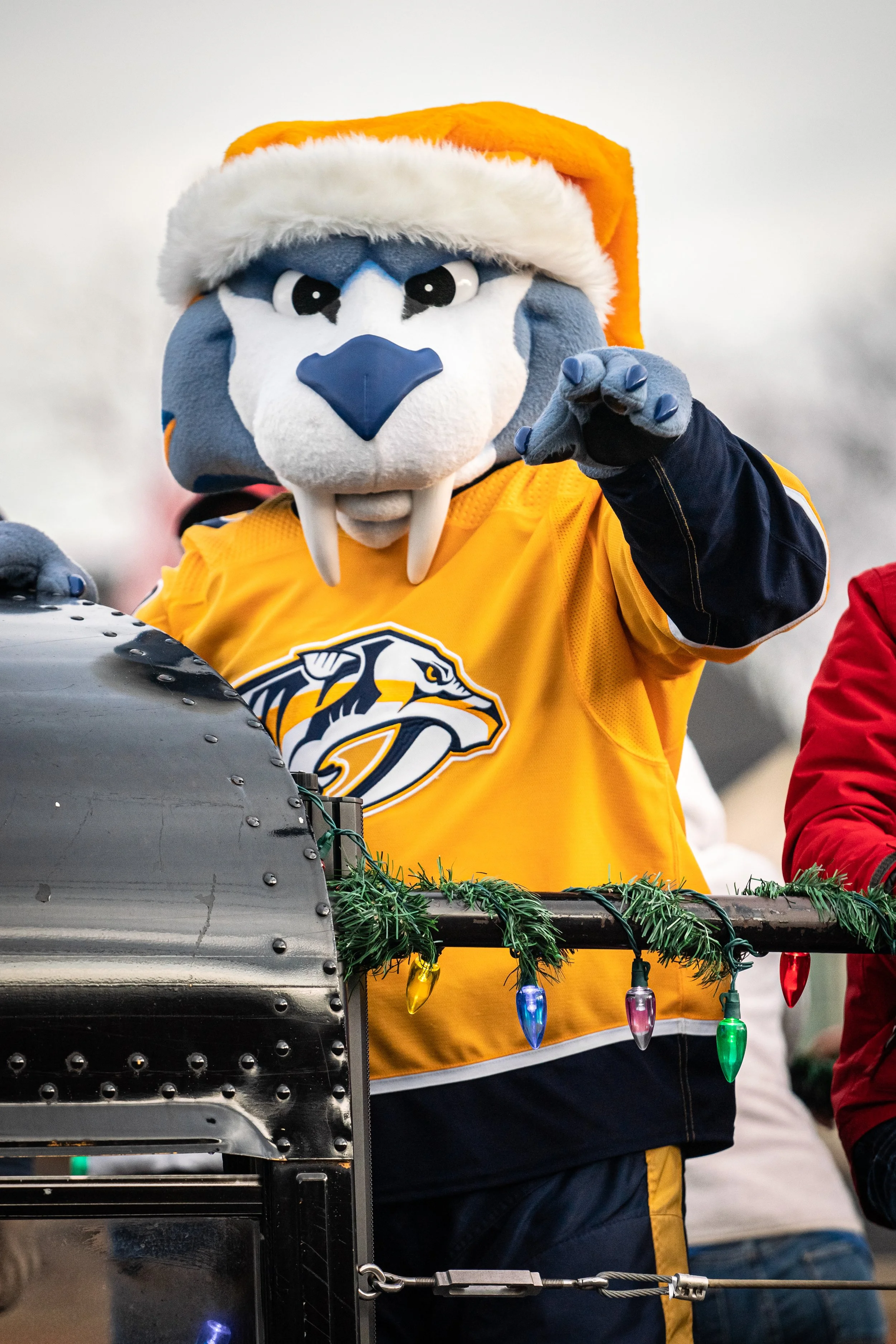 A person dressed in a lion mascot costume wearing a yellow sports jersey and an orange Santa hat, pointing towards the camera, standing behind a decorated and lit float with Christmas lights.
