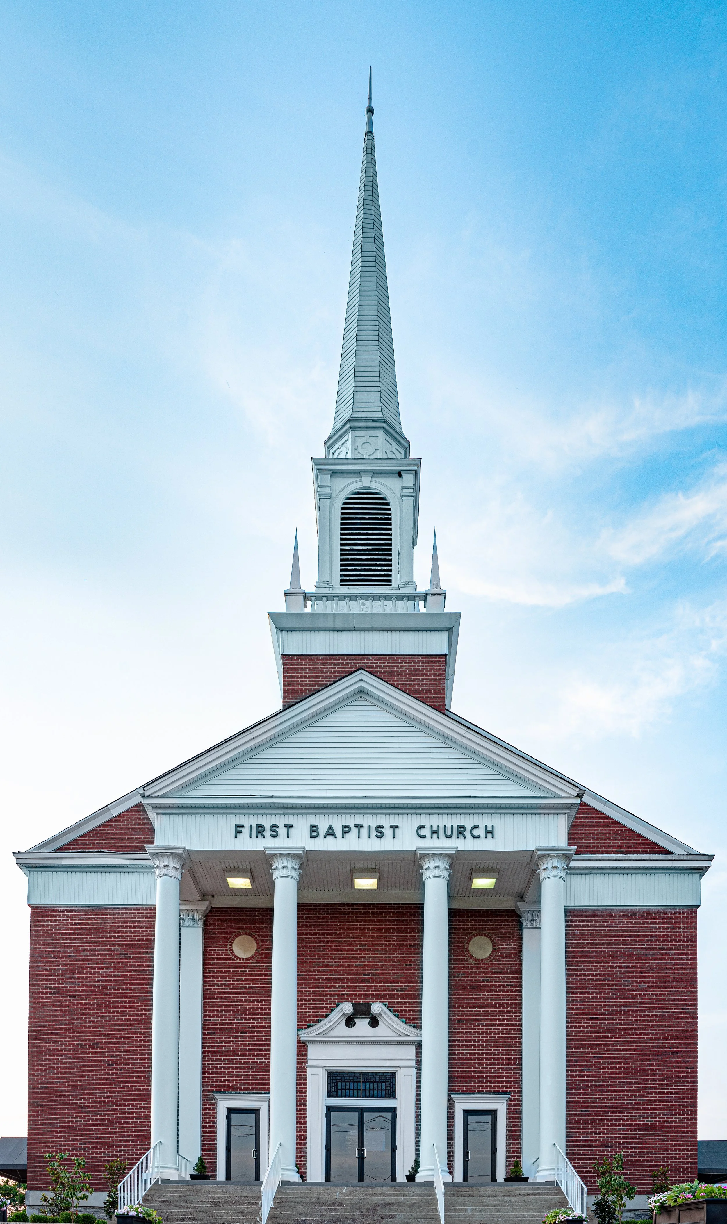 Front view of First Baptist Church with red brick facade, white columns, and tall steeple against a blue sky.