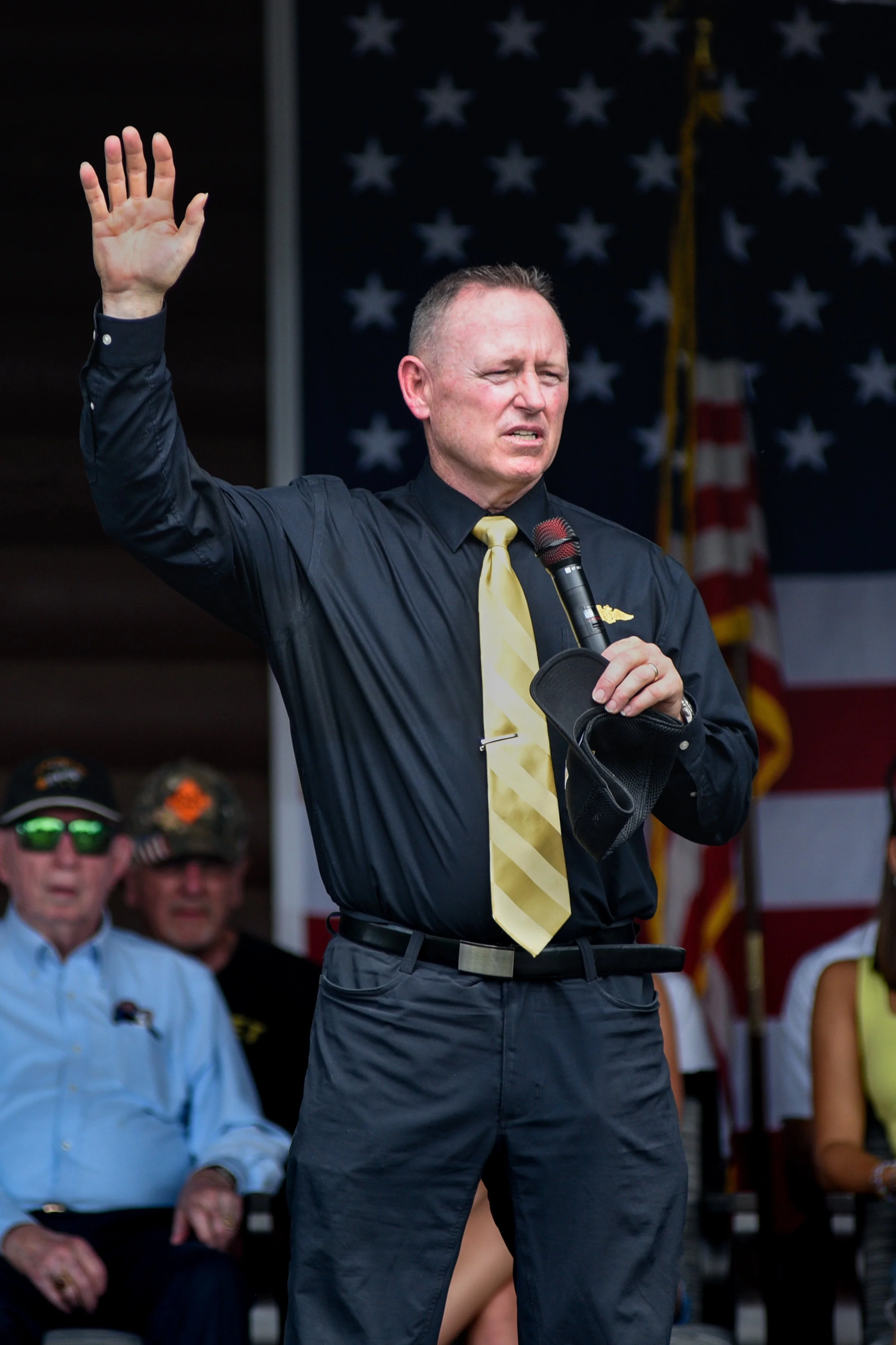 A man dressed in black with a yellow tie, holding a microphone and raising his hand, appears to be speaking at a public event or rally with an American flag in the background.