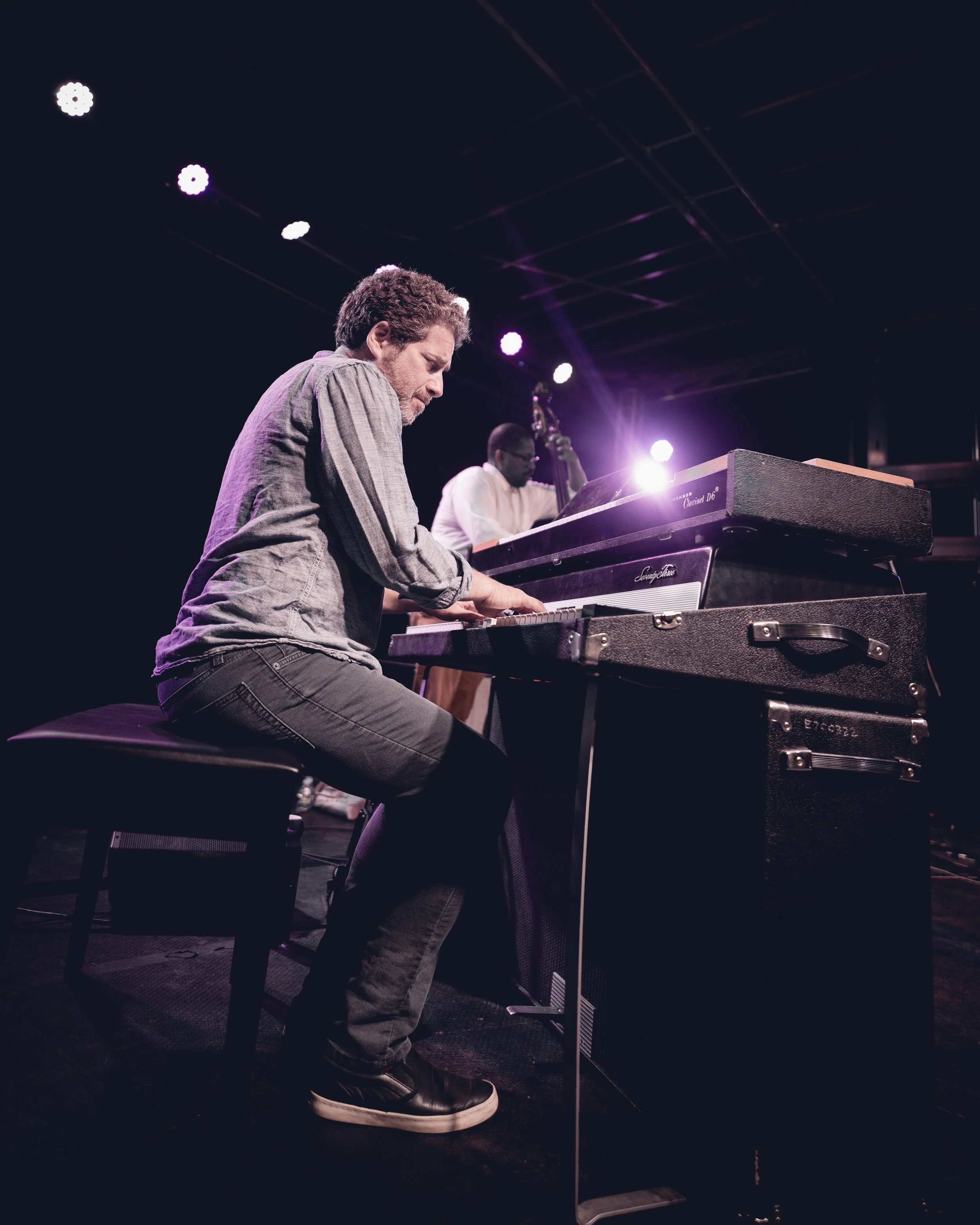 Man playing a grand piano on stage, purple and white stage lights overhead, dark background.