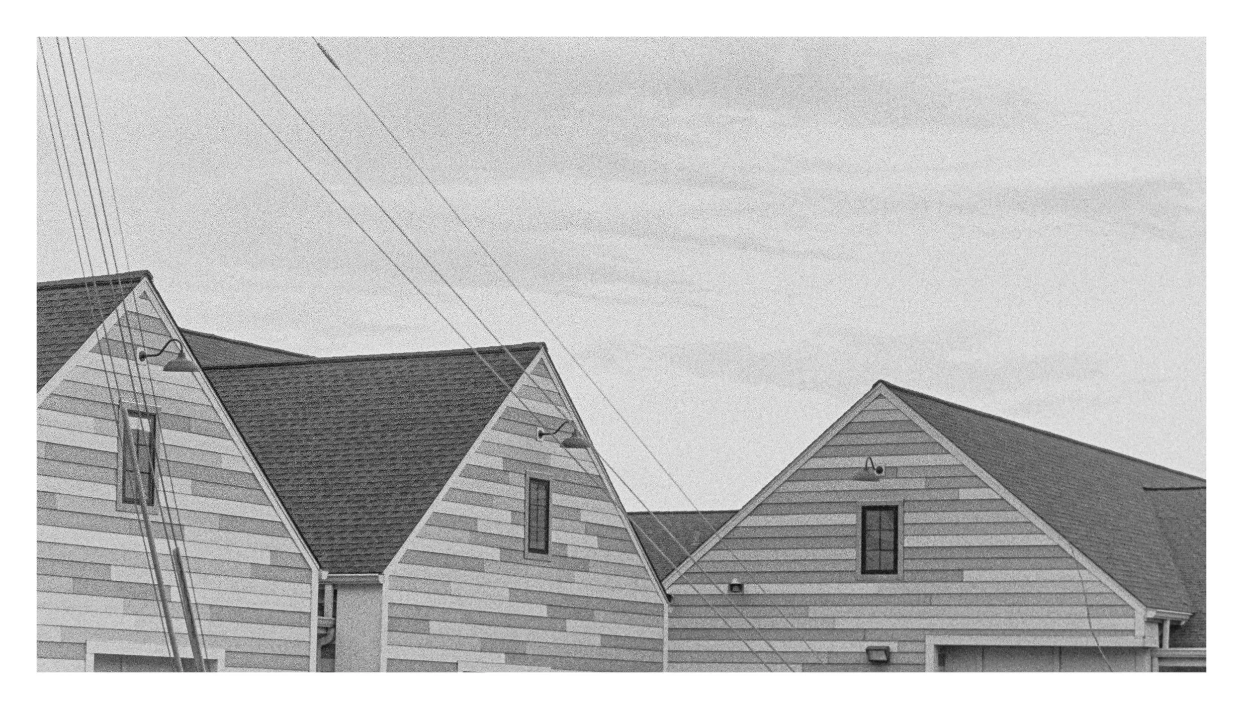 Black-and-white photo of three modern houses with peaked roofs, horizontal siding, and tall narrow windows, set against a sky with faint clouds.