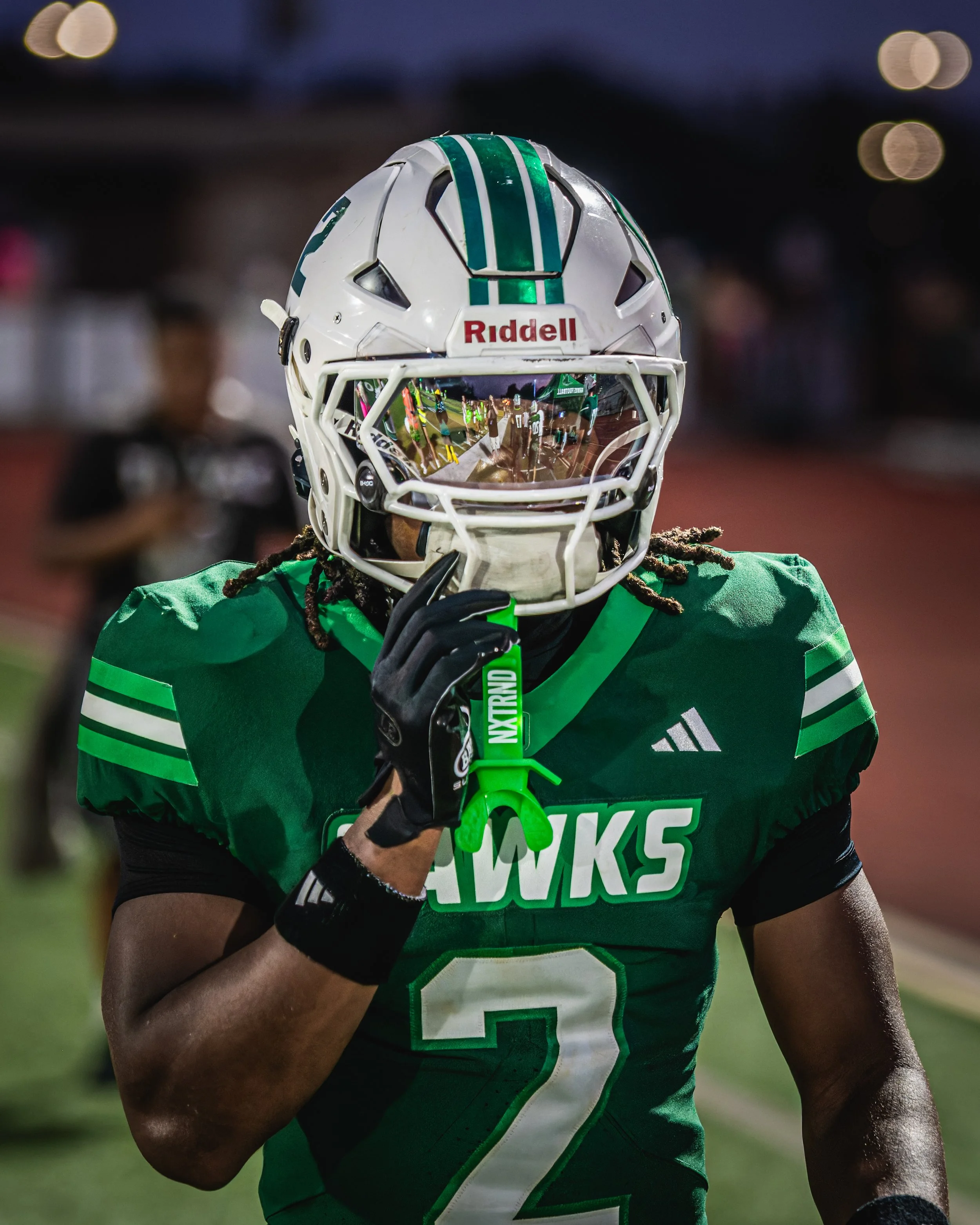 Close-up of an American football player wearing a green jersey and white helmet with green stripes, holding a green football, during a game at night.