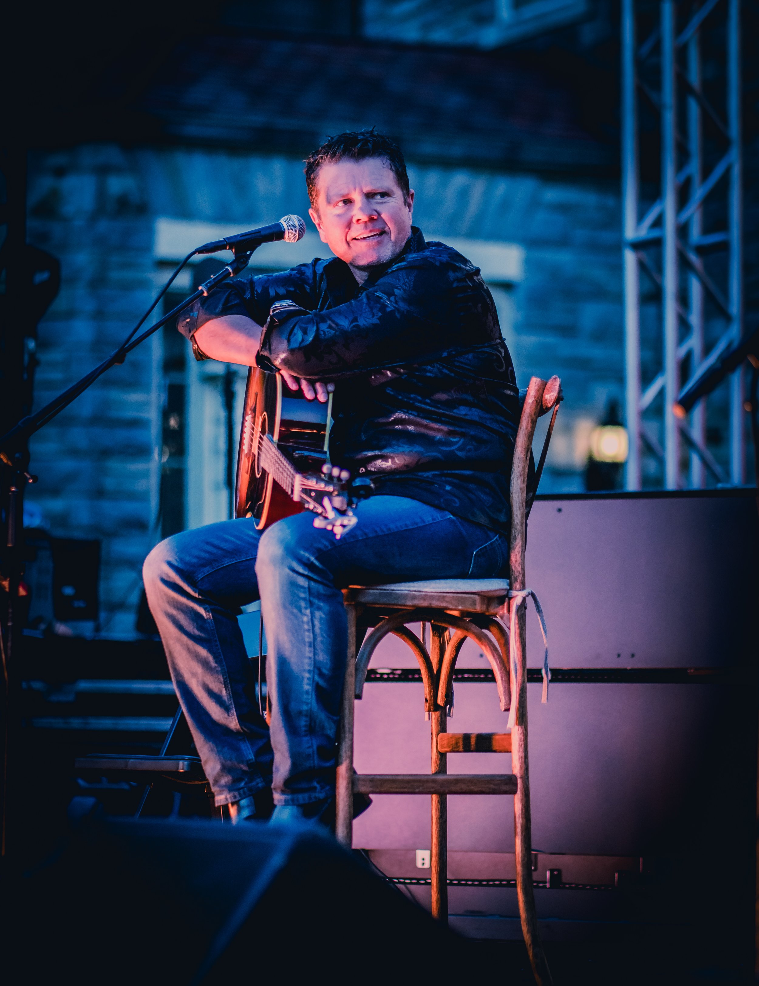A man sitting on a chair plays an acoustic guitar and sings into a microphone on a stage with blue lighting.