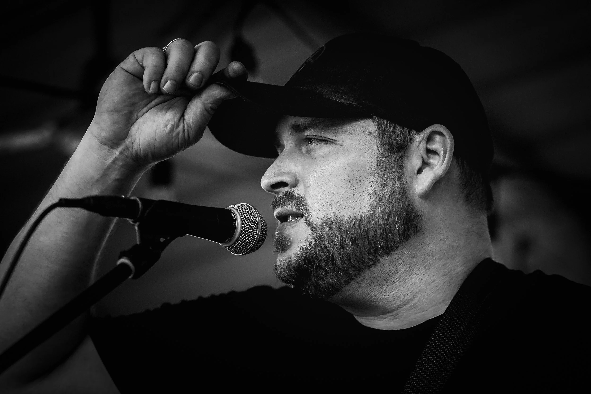 A black-and-white photo of a young man with a beard, wearing a dark cap and shirt, speaking into a microphone, with his hand adjusting his cap.