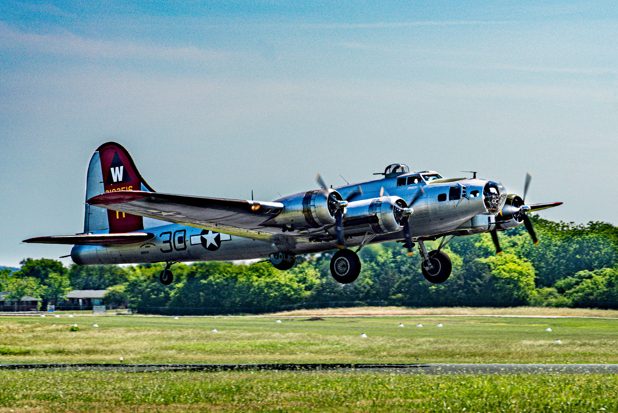 A vintage silver military aircraft in flight over a grassy field with trees in the background under a clear blue sky.