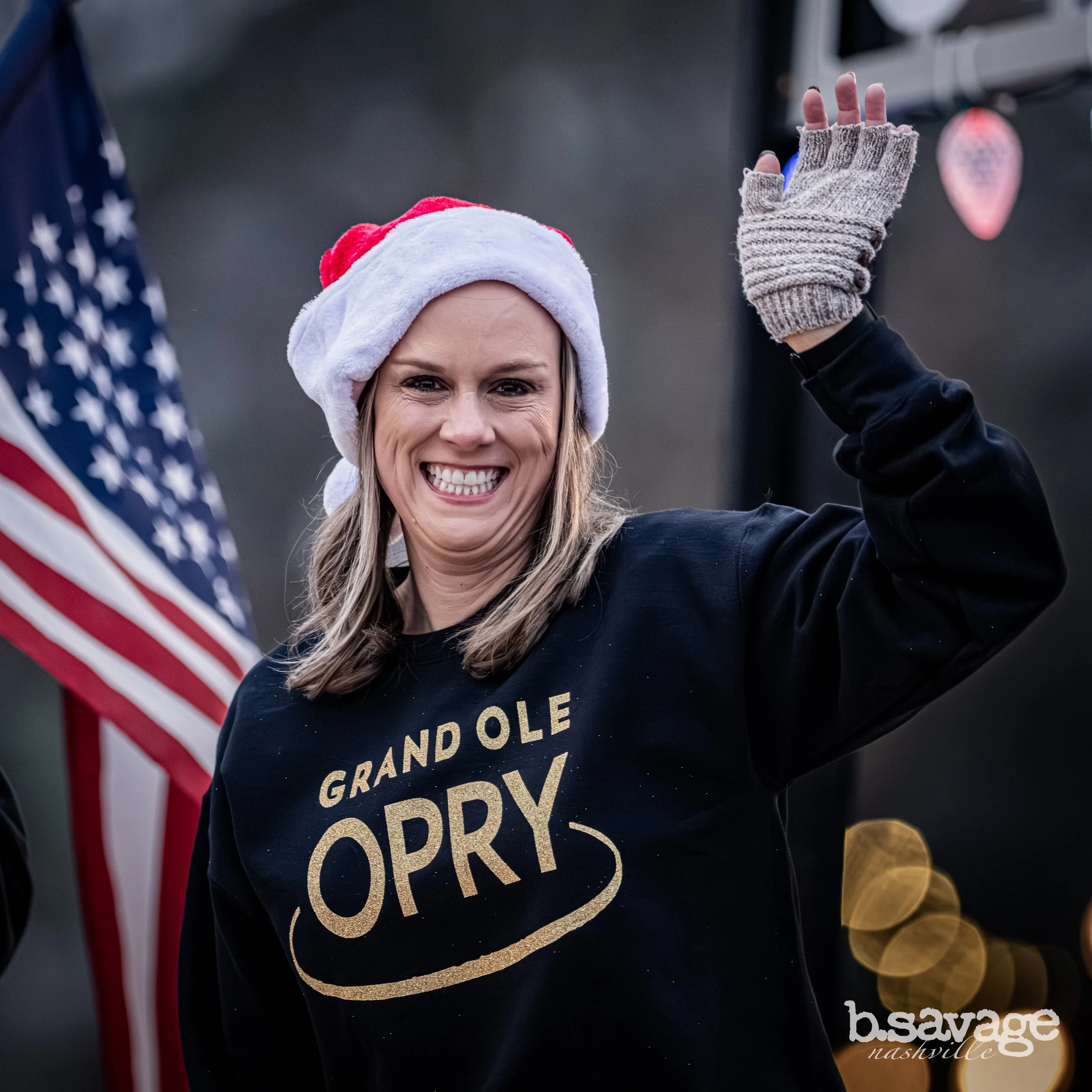 A woman smiling and waving, wearing a Santa hat and gray fingerless gloves, at a patriotic event with an American flag in the background, and gold coins on the ground.