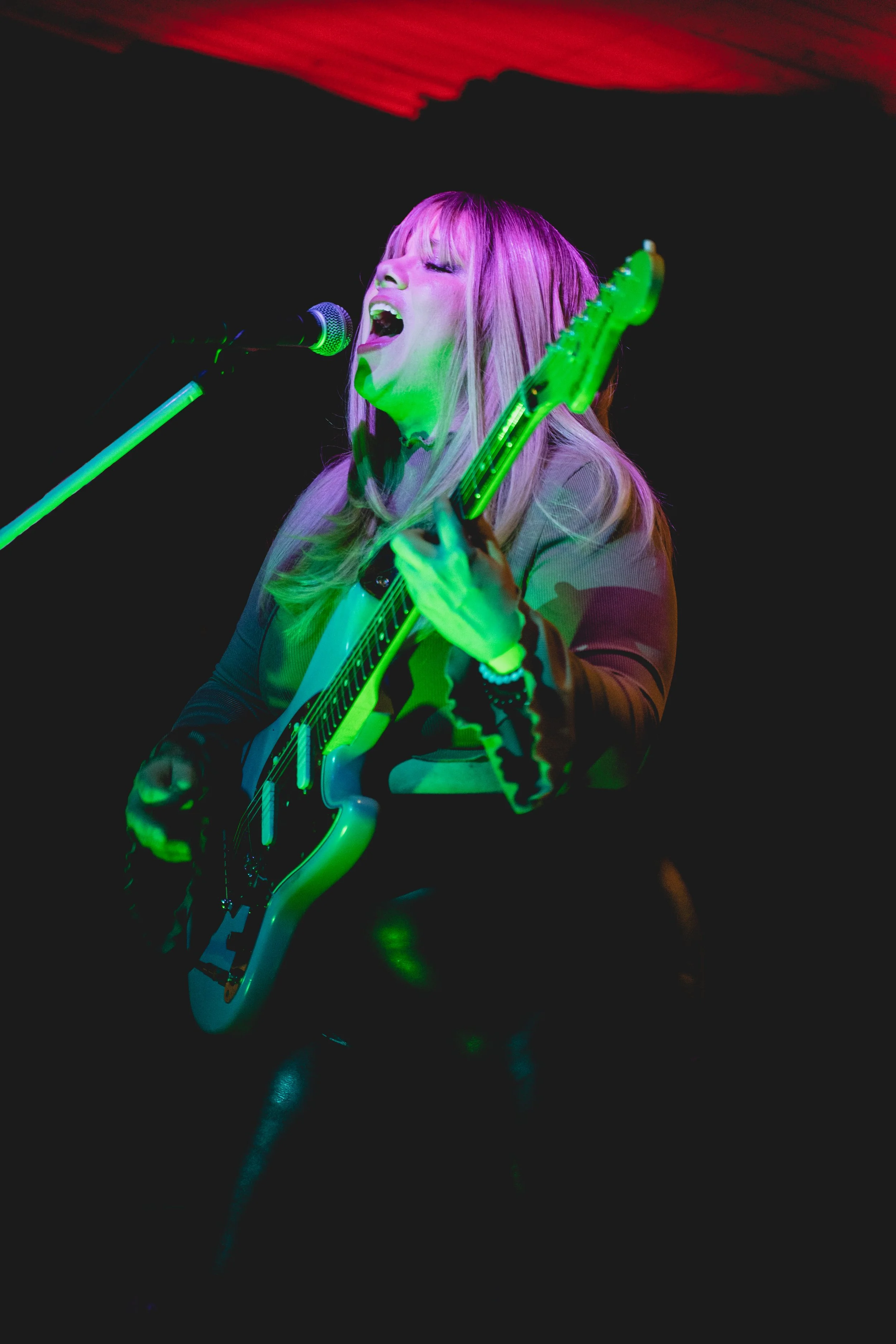A woman passionately singing and playing an electric guitar on stage, illuminated by colorful stage lights against a dark background.