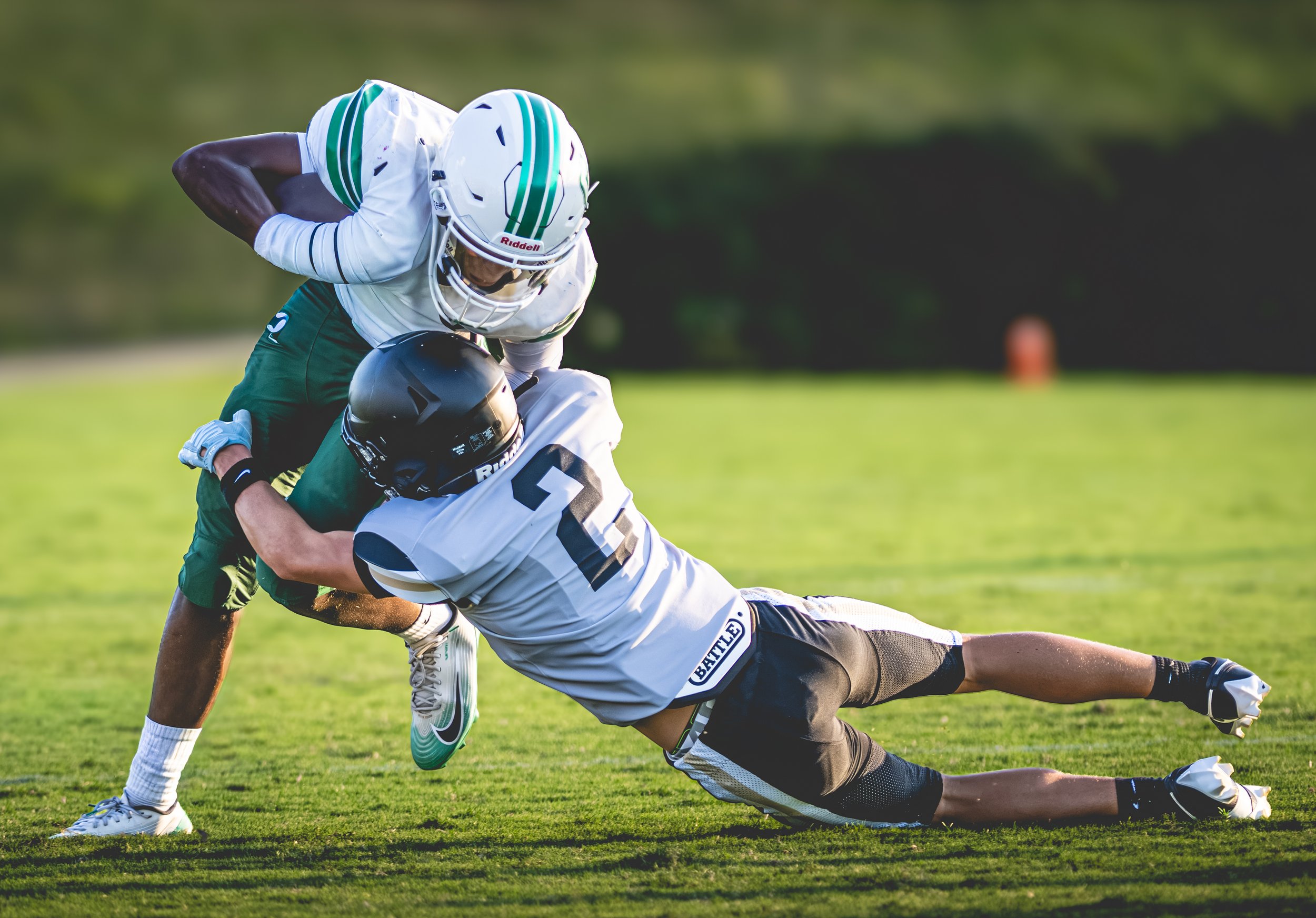 Two football players, one in a white uniform and another in a dark uniform, are engaged in a tackle on a grassy field during a game. The player in white is lunging and grabbing the player in dark uniform who is falling forward with his helmet close t
