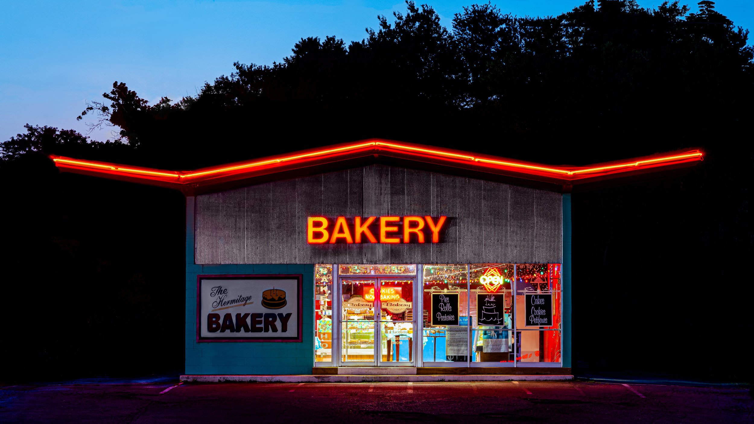 Nighttime view of a bakery storefront with red neon lights outlining the roof. The shop has a large digital sign reading 'BAKERY' in bright red. The entrance is lit with colorful neon signs advertising cookies, cakes, and pastries.