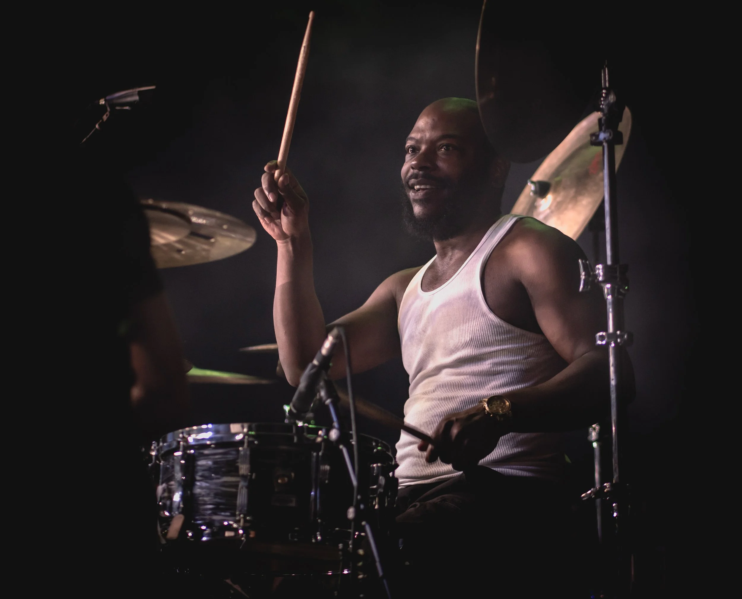 A man with a dark beard, wearing a white tank top and a gold watch, is playing drums in a dark setting.
