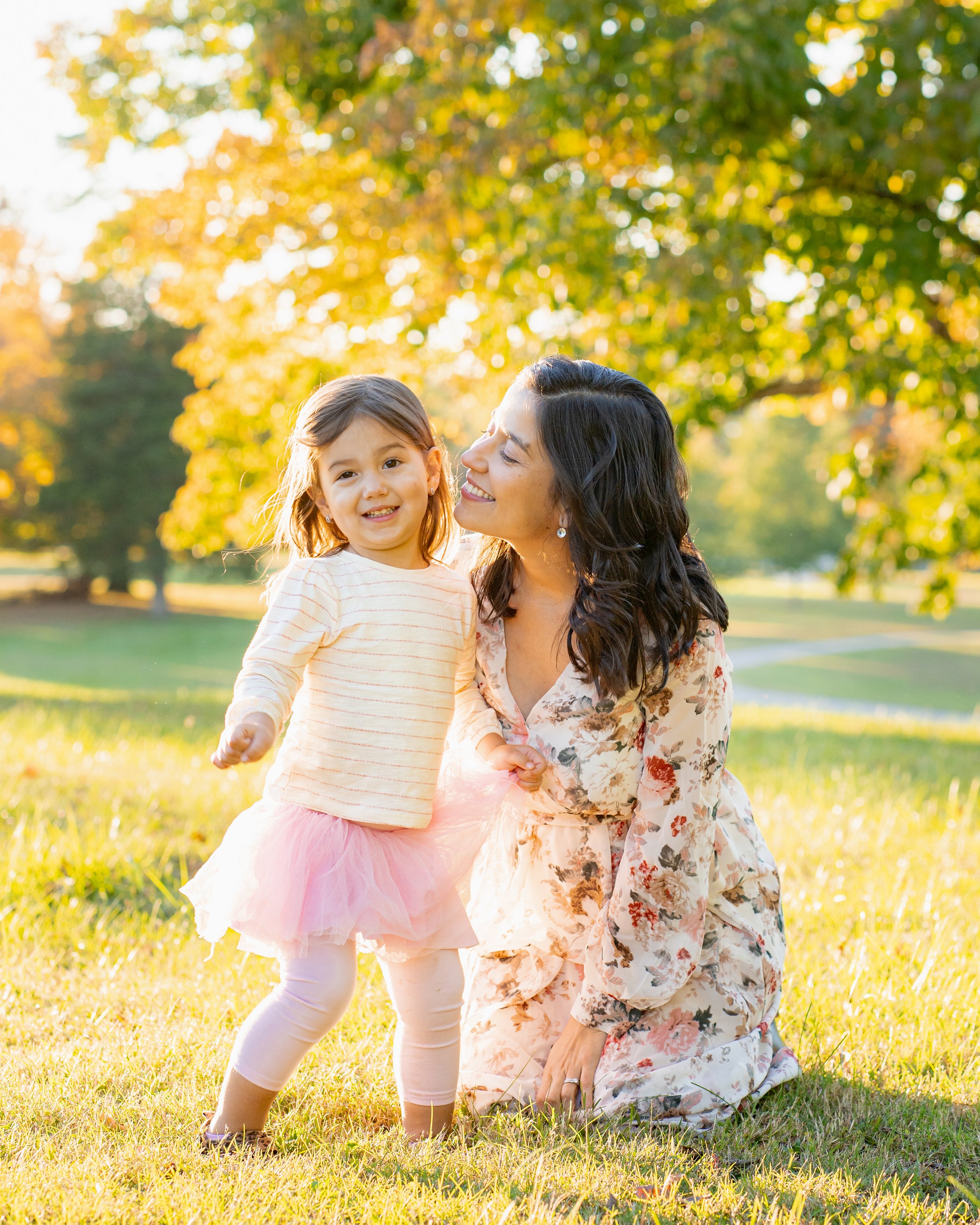 A woman and a young girl smiling outdoors in a park with yellow and green trees, bright sunlight, and grass, during autumn.