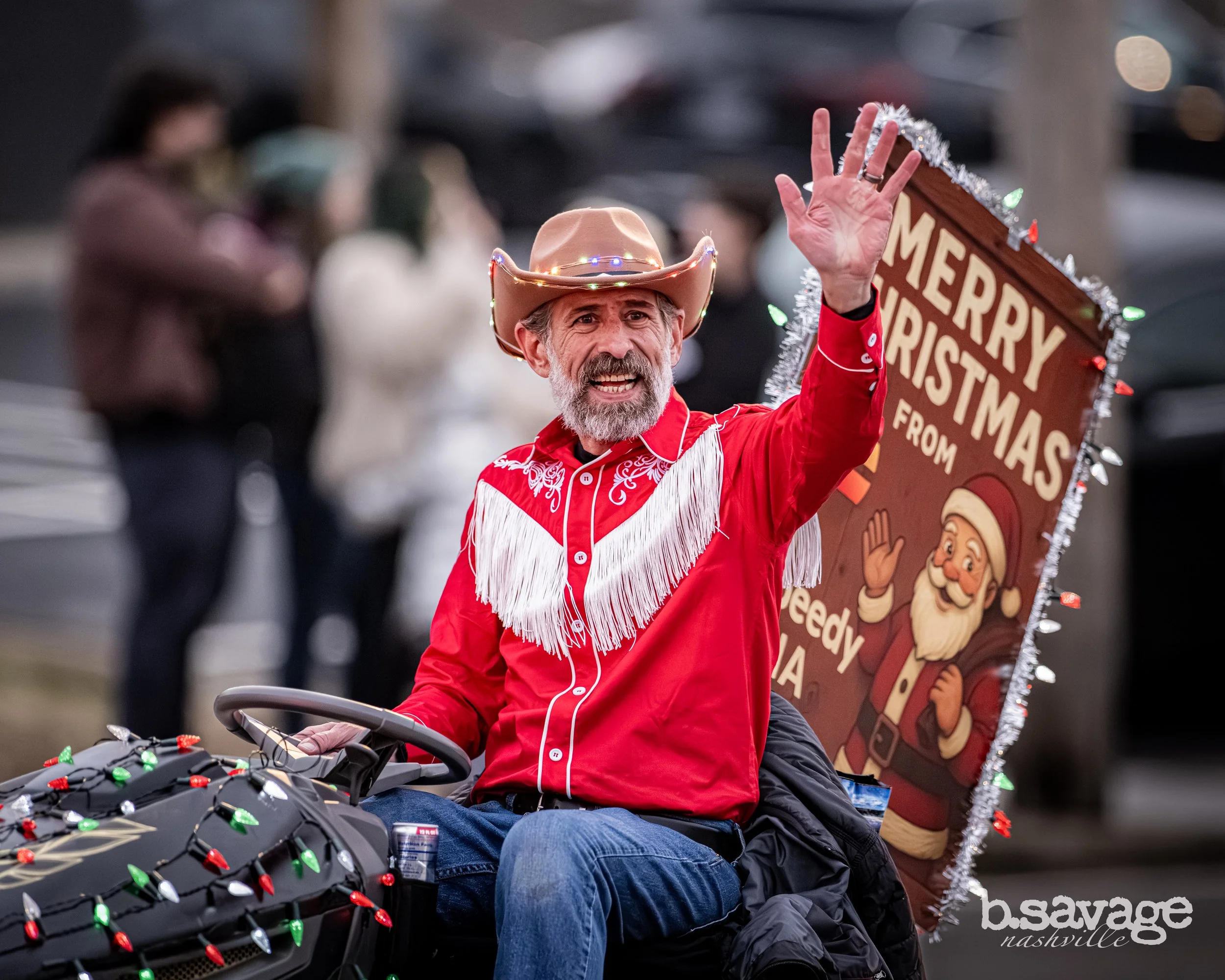 A man with a gray beard and light skin wearing a brown cowboy hat and a red shirt with white embroidery on the shoulders, sitting on a motorcycle decorated with red, green, and white Christmas lights. He is waving with his left hand and smiling. Ther