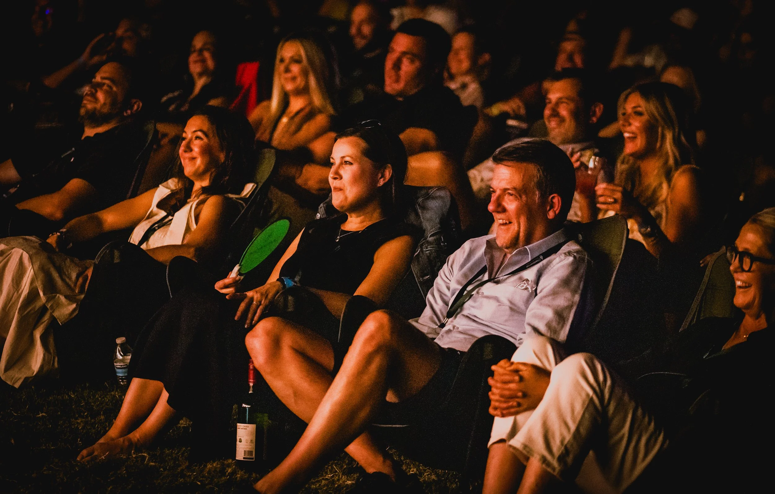 People sitting outdoors at night, laughing and enjoying a social event, some holding drinks and small fans.