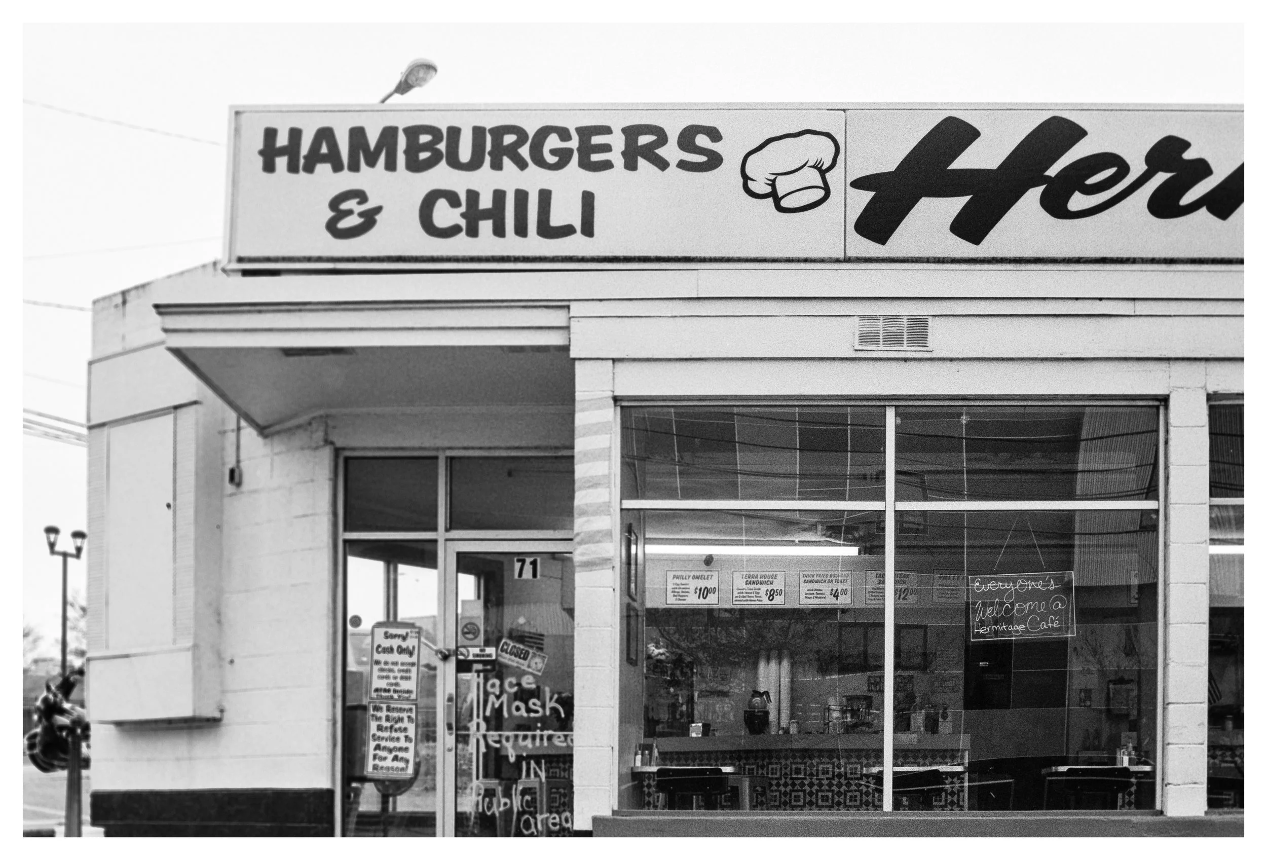 The storefront features a sign advertising hamburgers and chili with a chef's hat logo, with additional signs for foods like Philly omelet, terrarium house sandwich, and other menu items, and a chalkboard sign welcoming customers to Heritage Cafe.