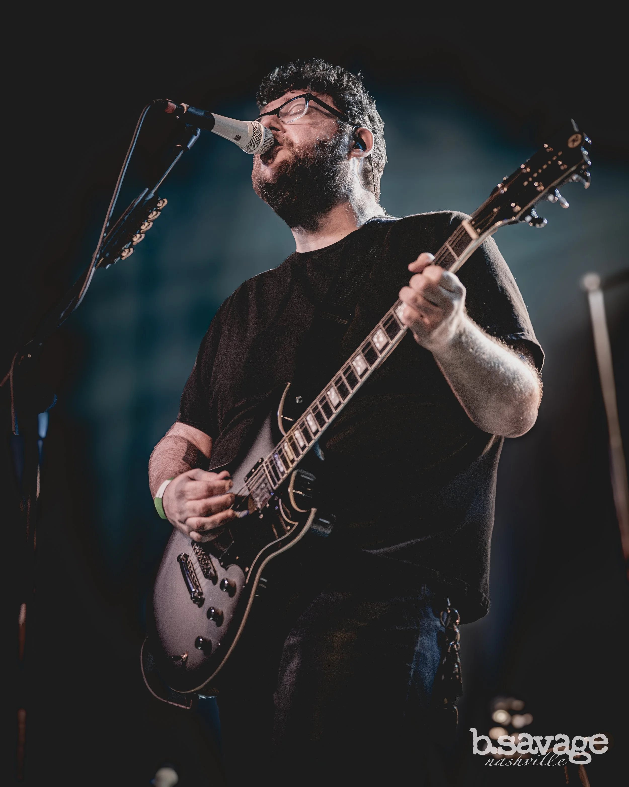 Male musician with curly hair, glasses, and beard singing into a microphone while playing an electric guitar on stage at a Nashville event, with the logo 'b.savage nashville' visible in the bottom right corner.