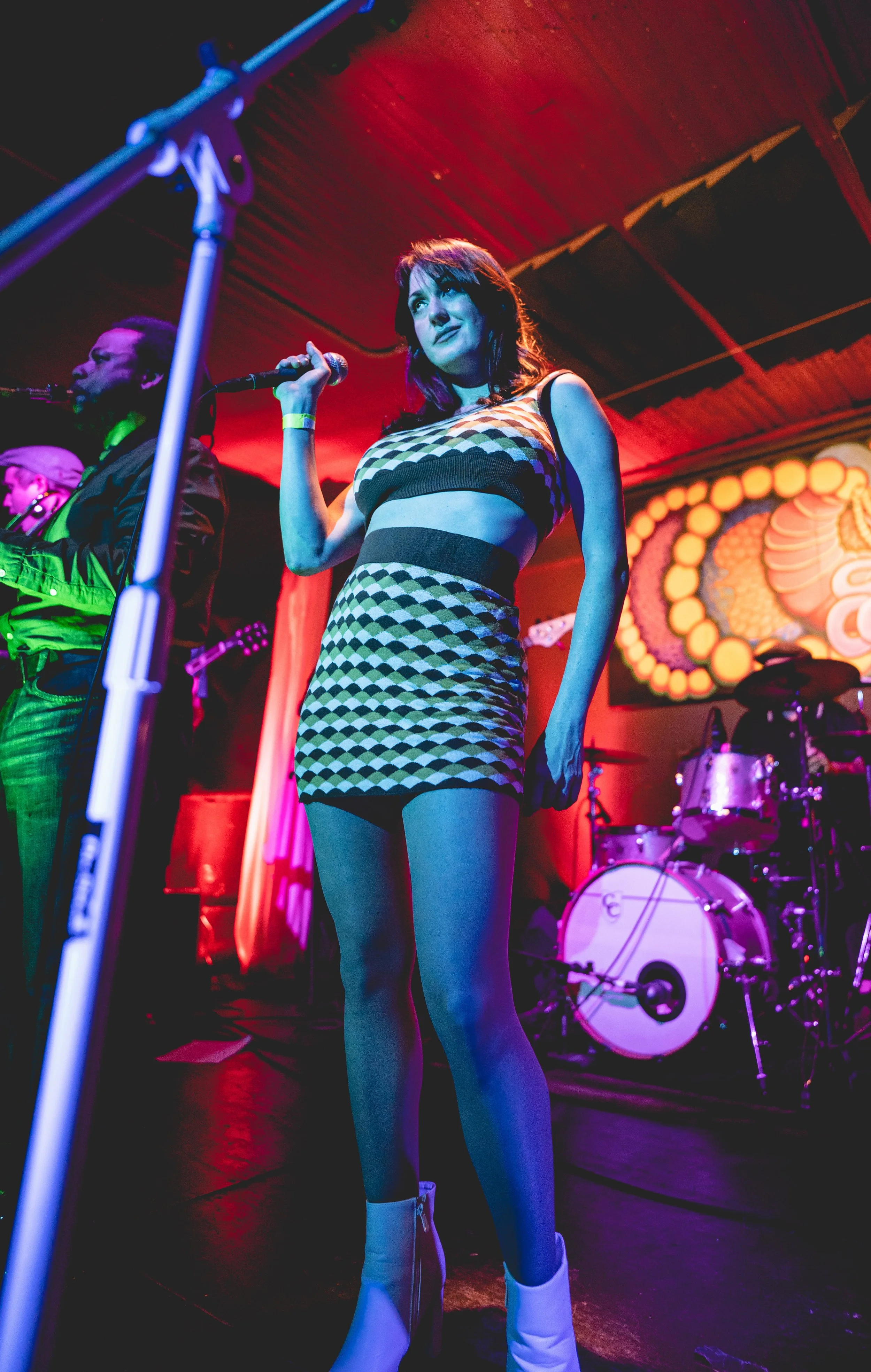 A woman with dark hair wearing a checkered crop top and matching skirt holding a microphone on stage with colorful lighting, with a band and drums in the background.