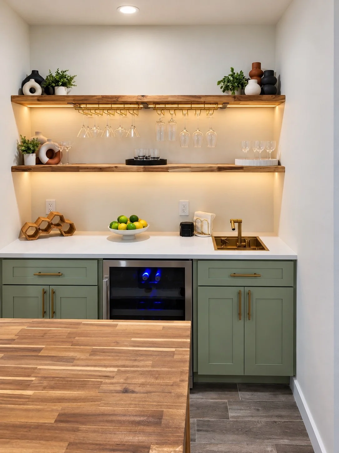 Sage green wet bar cabinetry with gold hardware and illuminated wood shelves.