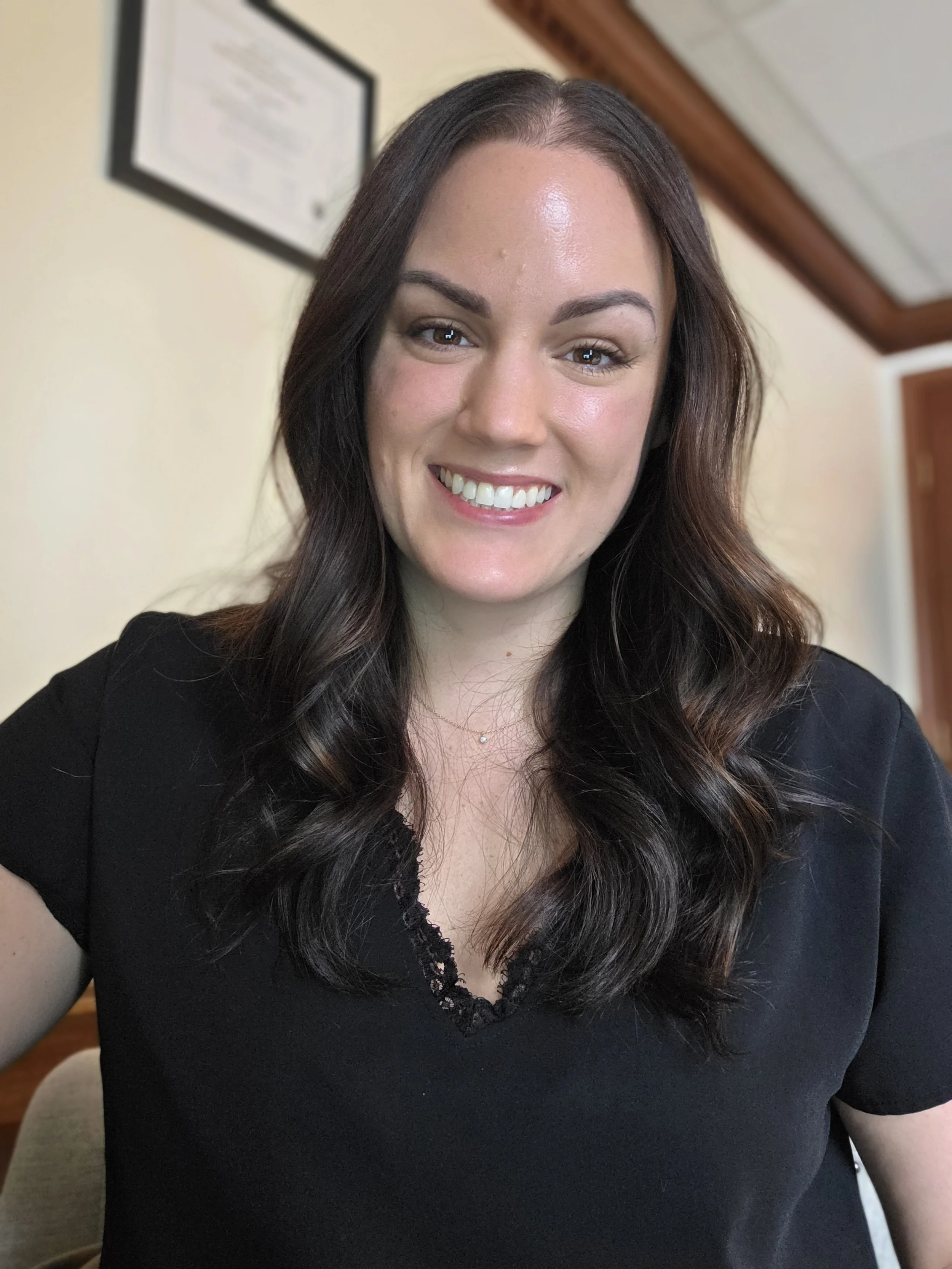 A woman with dark brown hair styled in loose waves, smiling with teeth showing, wearing a black top.