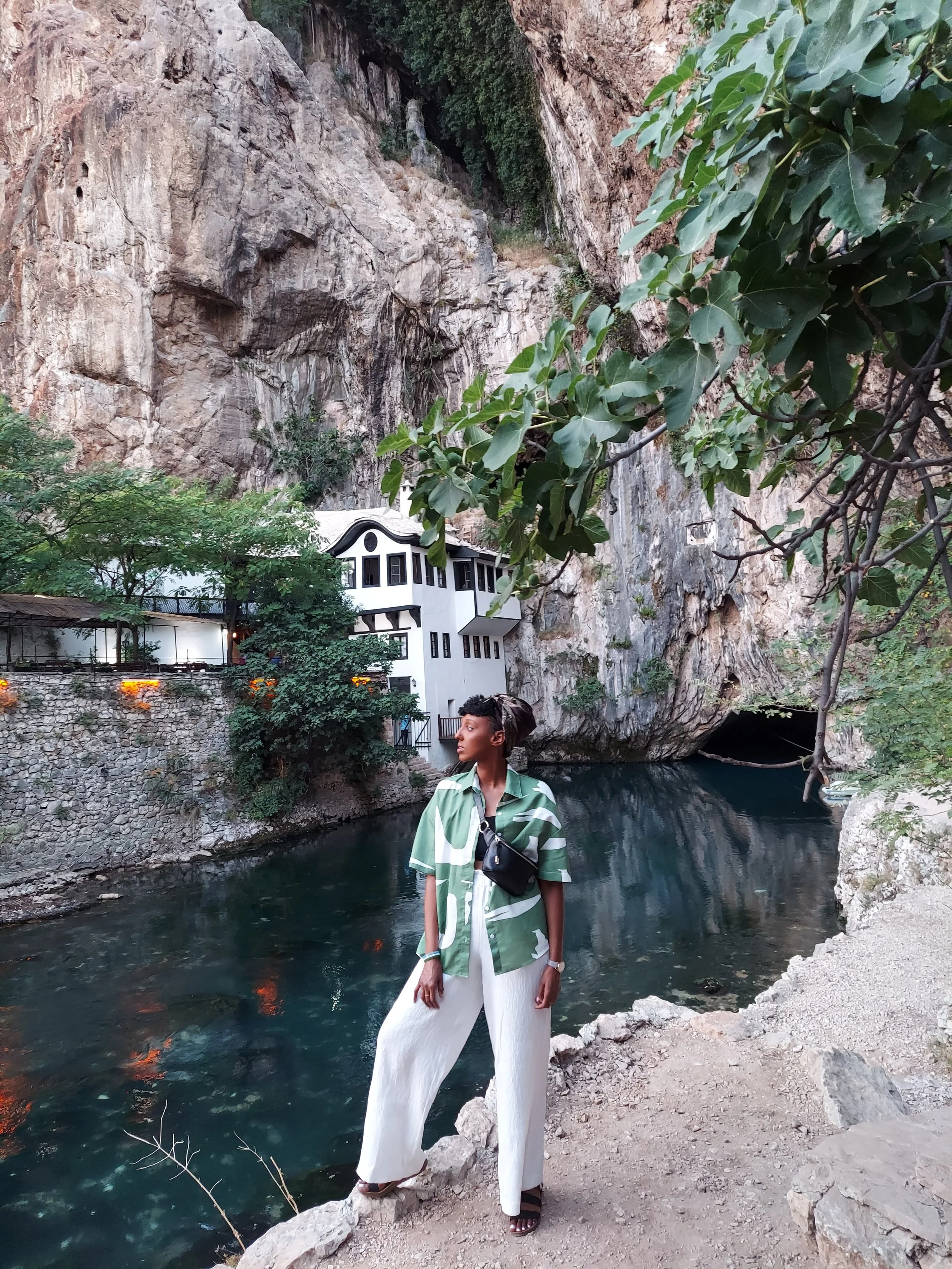 A woman standing near a river in front of a rocky cliff with buildings visible on the side of the cliff. She is wearing a green patterned shirt, white wide-leg pants, and sandals.