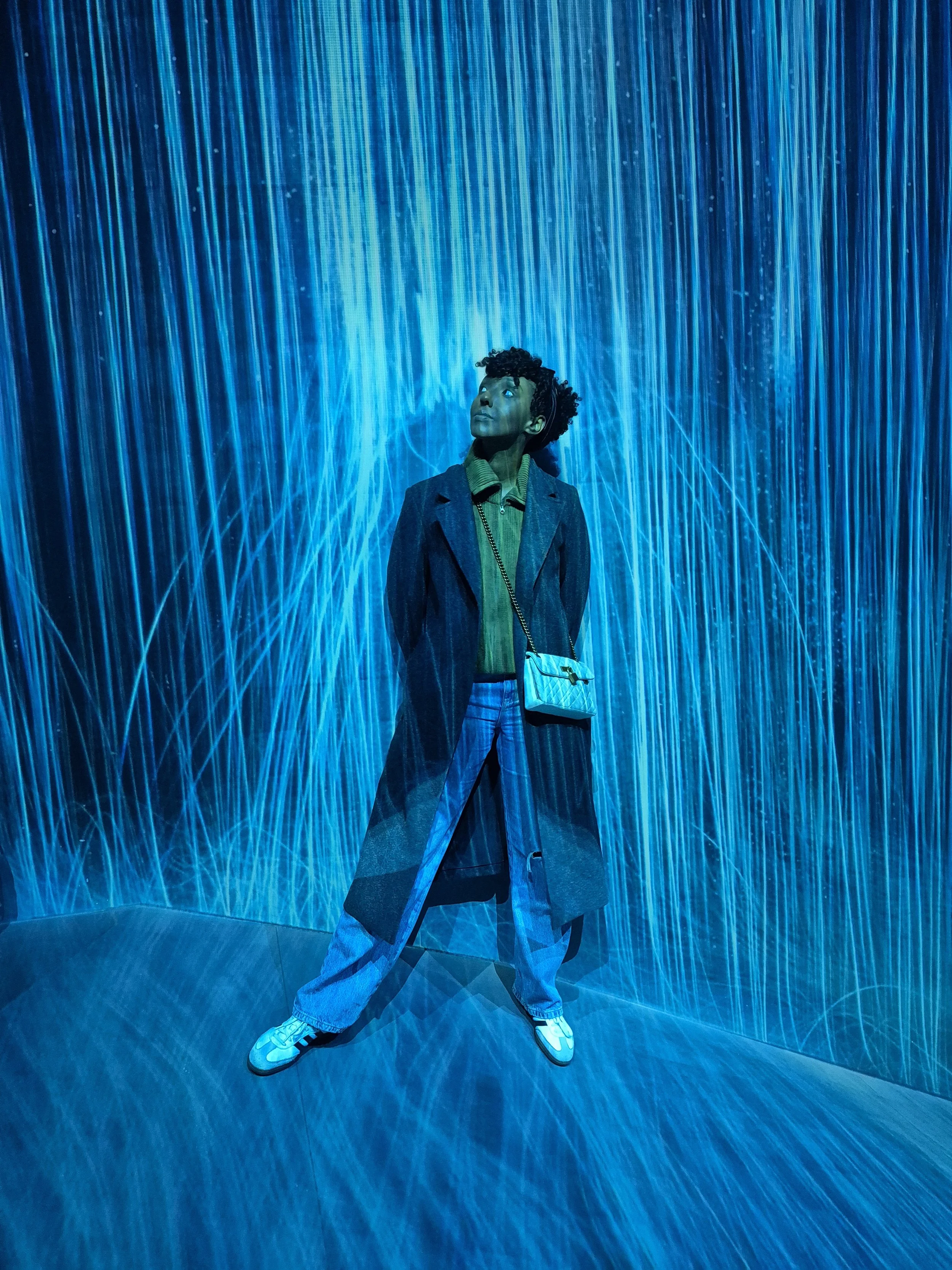 A person wearing a grey coat, green shirt, jeans, and sneakers, with a small crossbody bag, standing in front of a blue LED light installation with vertical light streams and fiber optic effects, looking up thoughtfully.