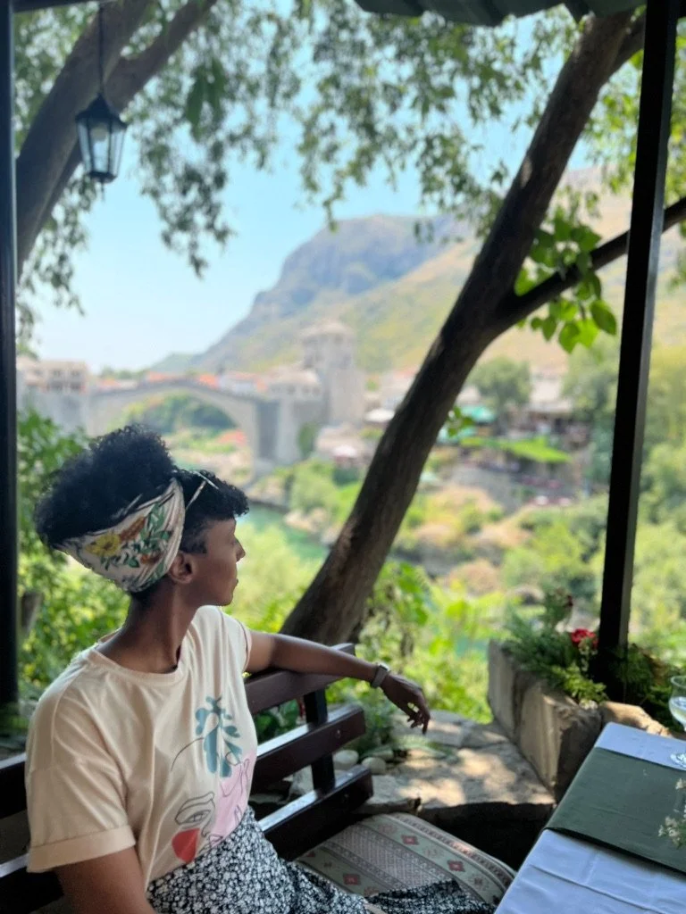 A woman sitting on a wooden bench outdoors, looking towards a scenic landscape with a bridge, a tower, and a hill in the background, surrounded by trees and greenery.