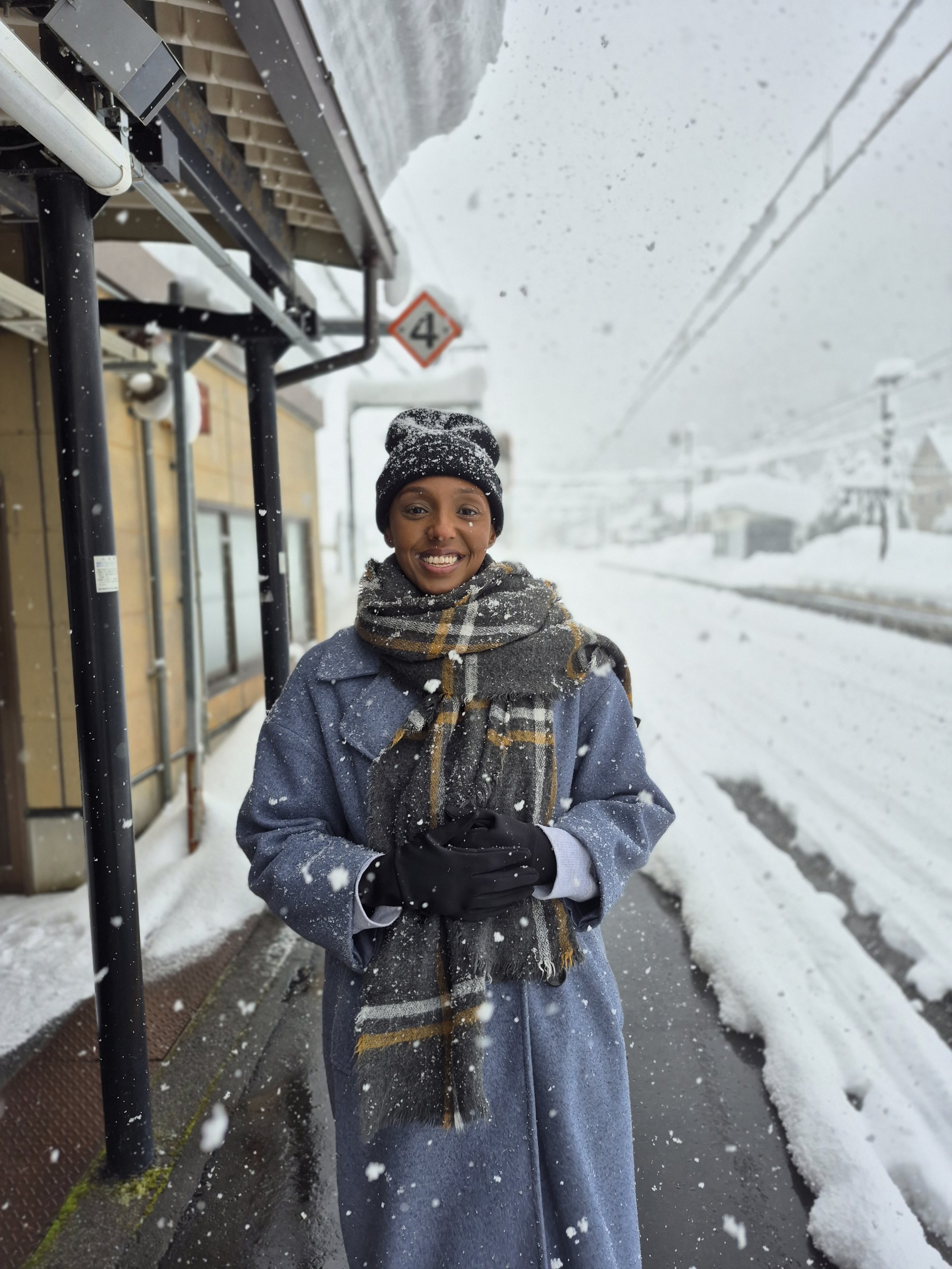 A smiling woman dressed warmly in a blue coat, plaid scarf, black gloves, and a black beanie, standing outside in snowy weather near a building and railroad tracks.