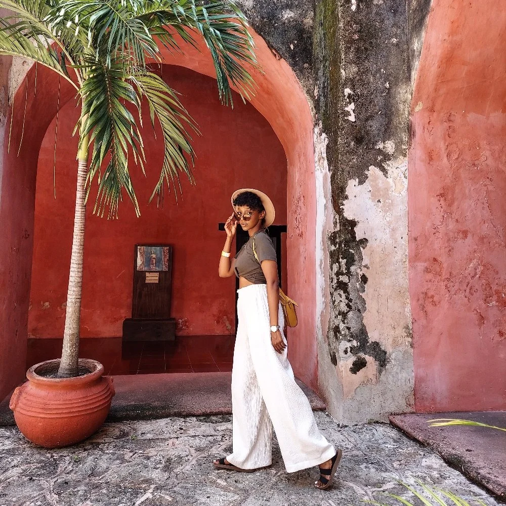 A woman stands in a courtyard next to a large potted palm tree, wearing wide-leg white pants, a brown crop top, a hat, sunglasses, and sandals, with a yellow purse over her shoulder.