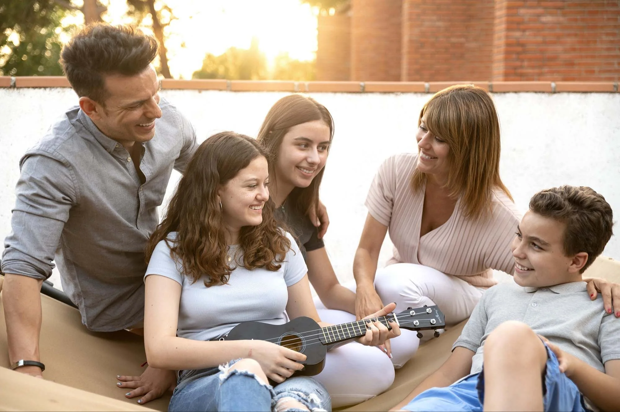 A group of six people, four women and two men, happily sitting outside on a sunny day. One woman is playing a small black ukulele while the others watch and smile. They are sitting on a beige outdoor sofa with a brick ledge and trees in the background.