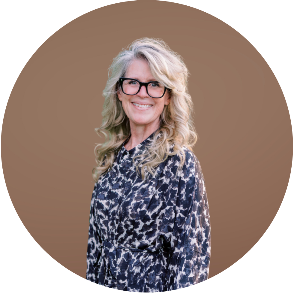 A woman with long curly blonde hair, black glasses, and a patterned dress, smiling at the camera against a tan background.