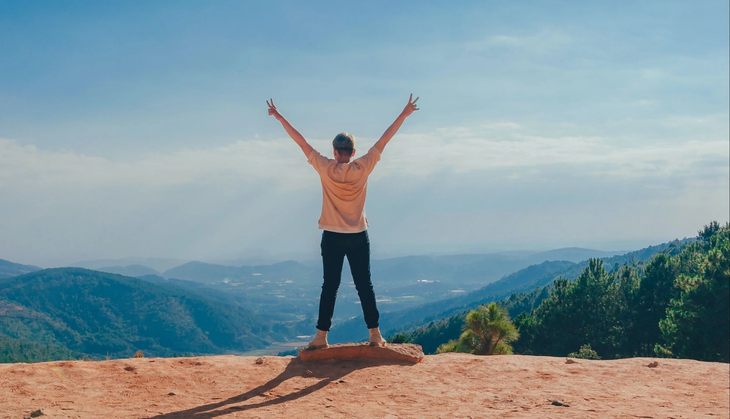 Person standing on a rock overlooking a valley with hills and trees, arms raised with peace signs, in a mountainous landscape under a partly cloudy sky.