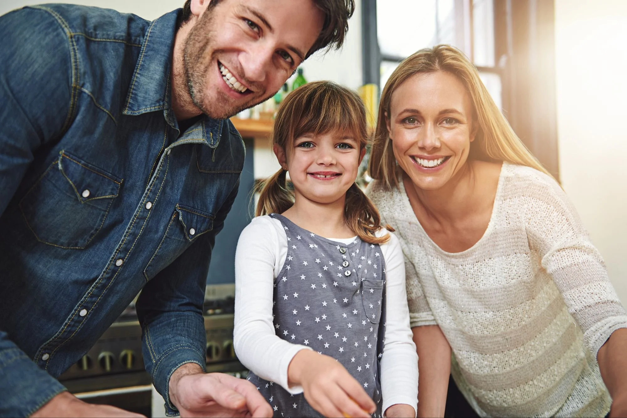 A smiling family of three, including a man, a woman, and a young girl, posing together indoors with sunlight streaming in, all showing happiness.