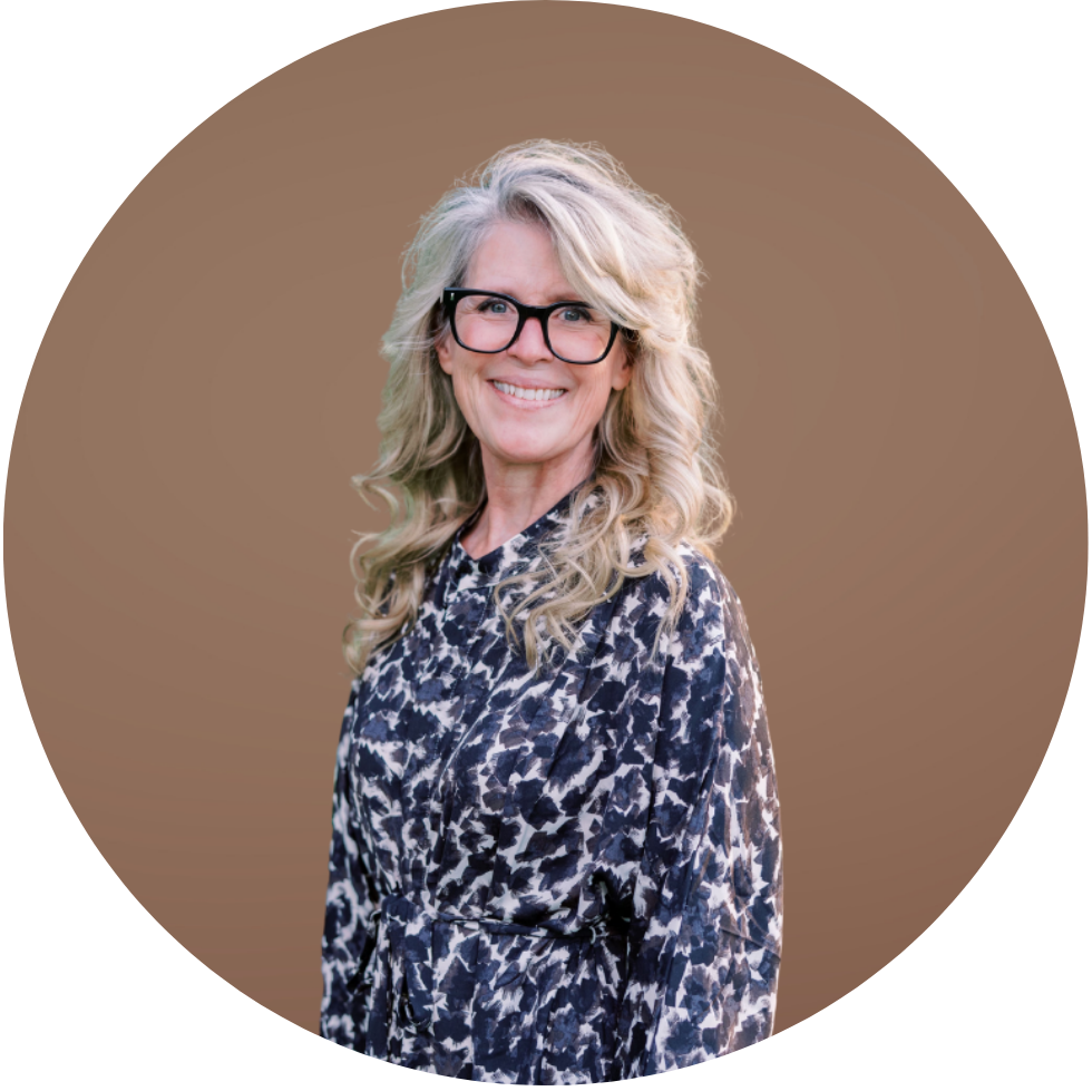 A smiling woman with long, wavy blonde hair and black glasses, wearing a black and white patterned blouse, standing against a brown background.