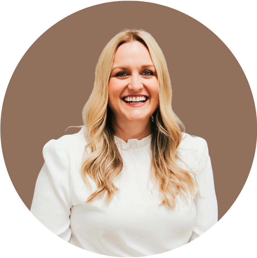 A smiling woman with long blonde hair, wearing a white blouse, standing against a tan background.