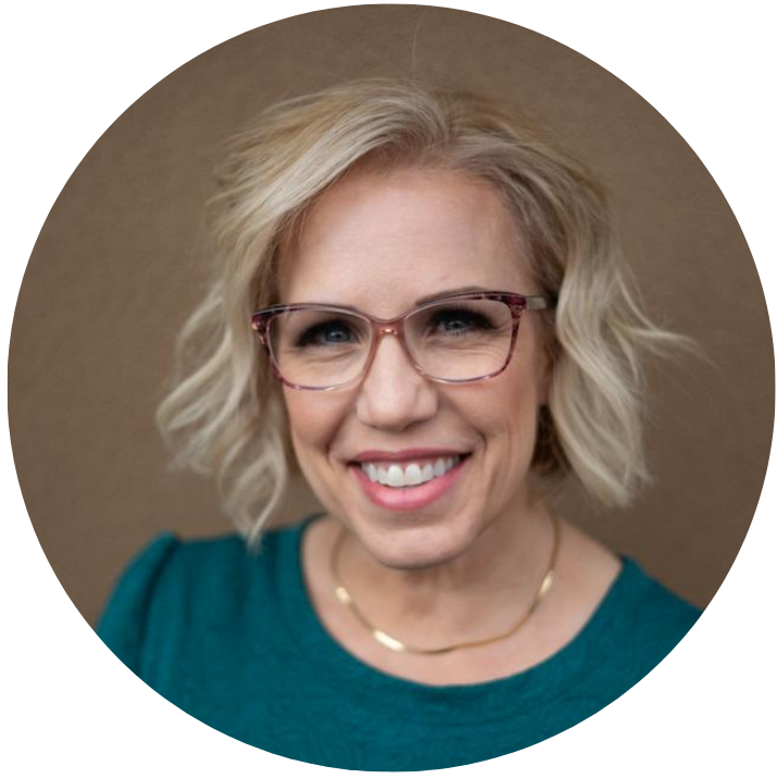 Close-up portrait of a smiling woman with blonde wavy hair, wearing glasses and a teal top, against a brown background.