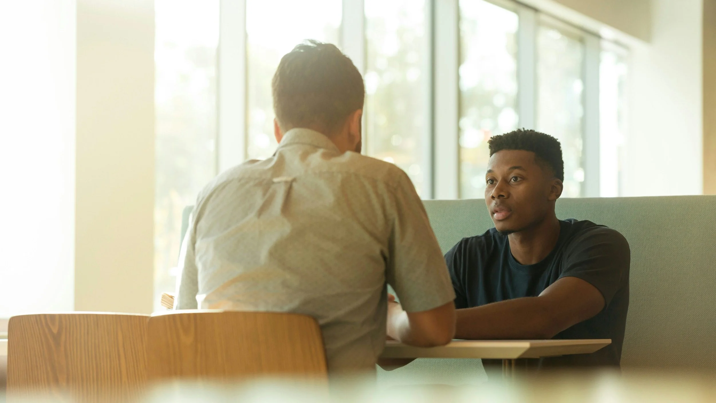 Two men having a conversation in a bright, indoor setting with large windows in the background.