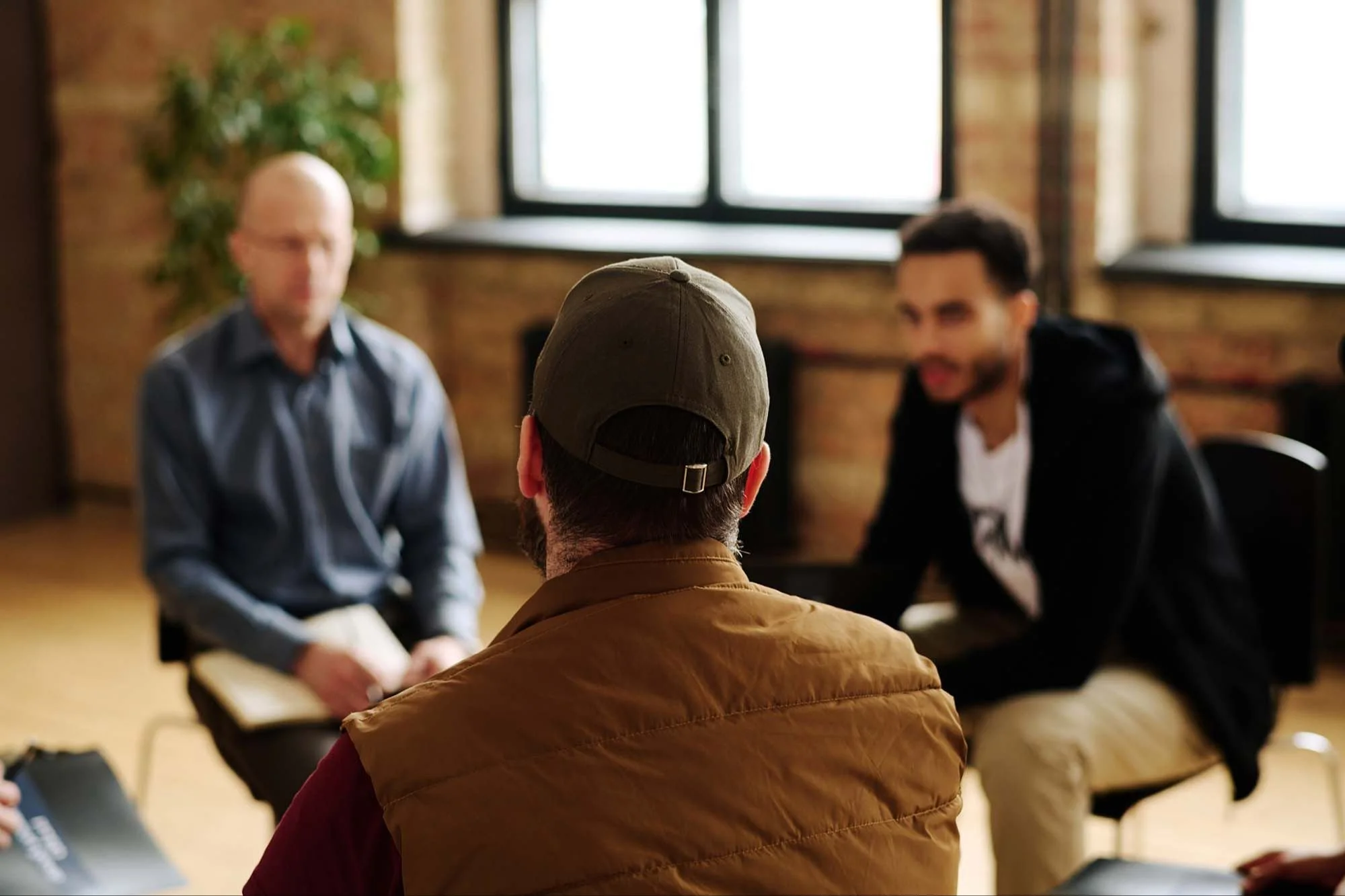 A man wearing a brown jacket and cap is sitting in front of a group of people in a room with brick walls and large windows, possibly during an interview or counseling session.