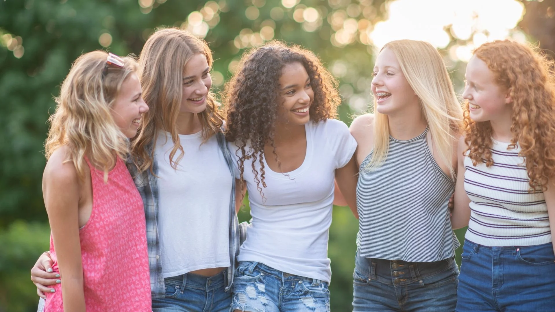 A group of five young women standing together outdoors, smiling and talking with each other.