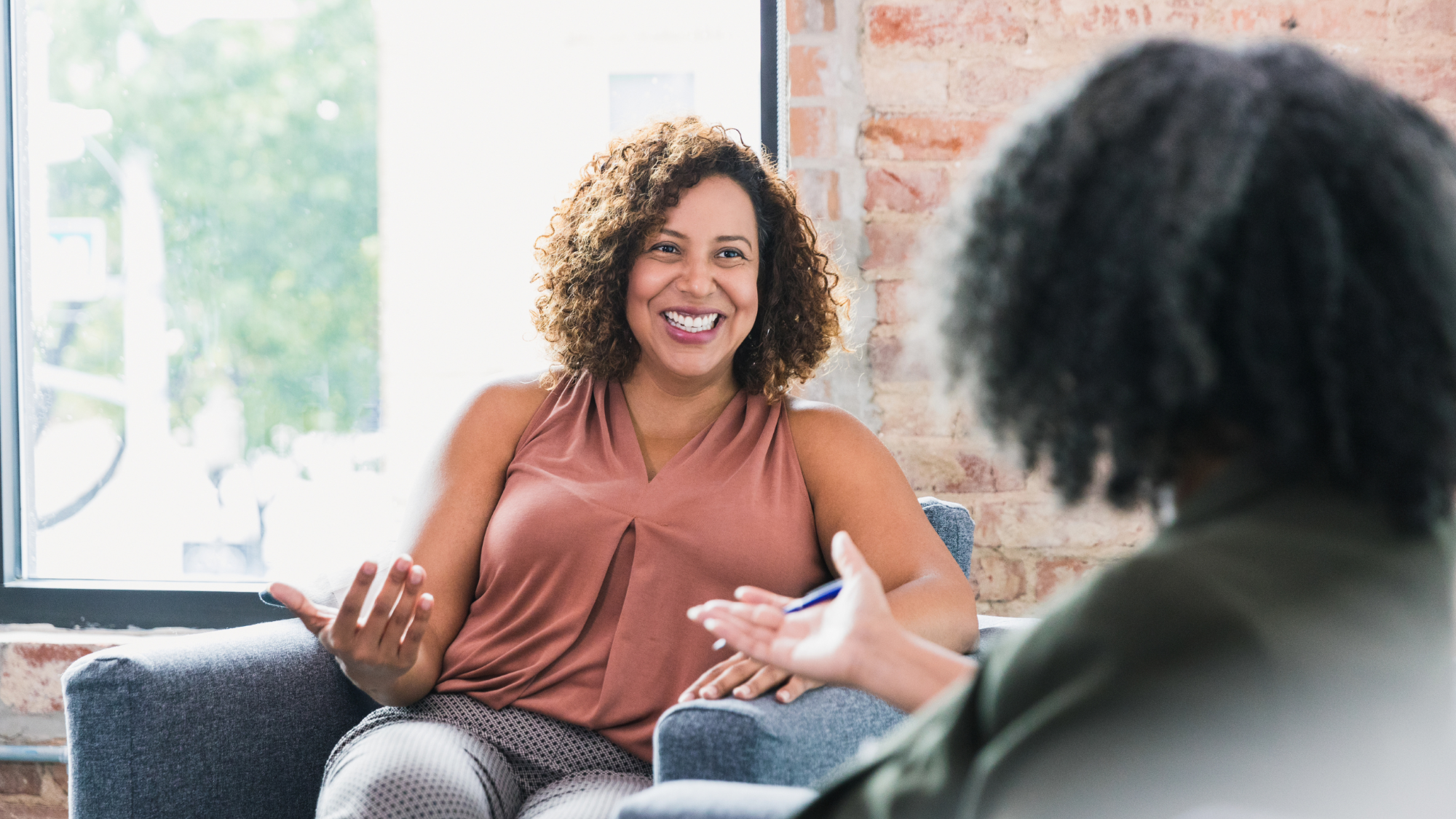 Two women having a conversation in a bright room with large windows and a brick wall. One woman, with curly hair wearing a sleeveless top, is smiling and gesturing with her hands. The other woman, with darker hair, is partially visible and engaged in the discussion.