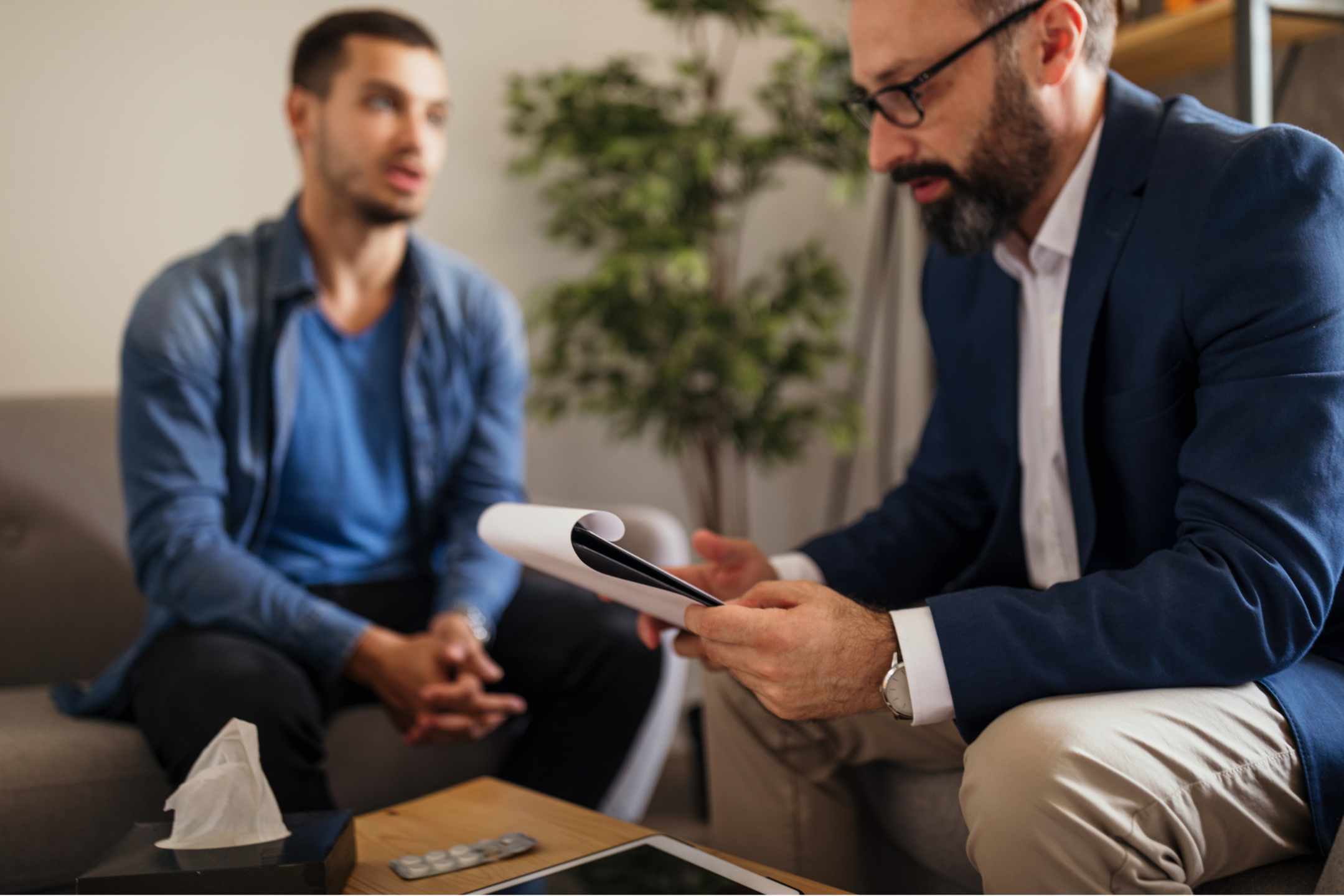 Two men in a meeting or counseling session, one holding papers, with a tissue box, tablets, and a smartphone on the table.