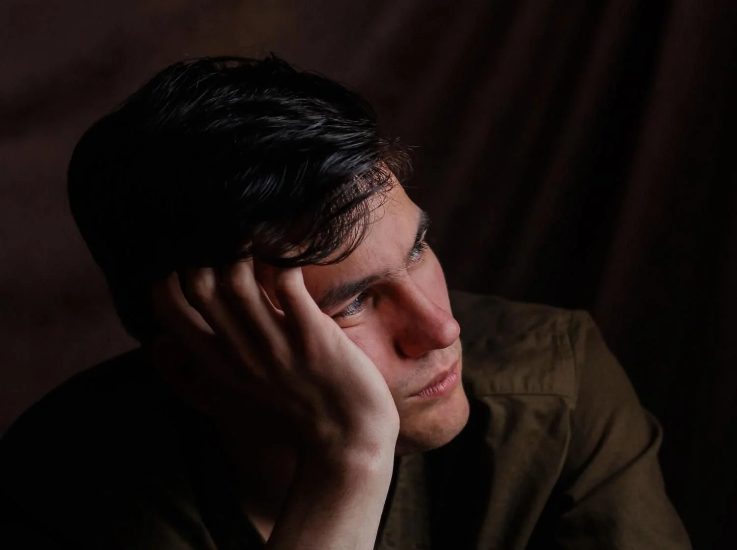 A young man with dark hair, resting his face in his hand, looking contemplative against a dark background.