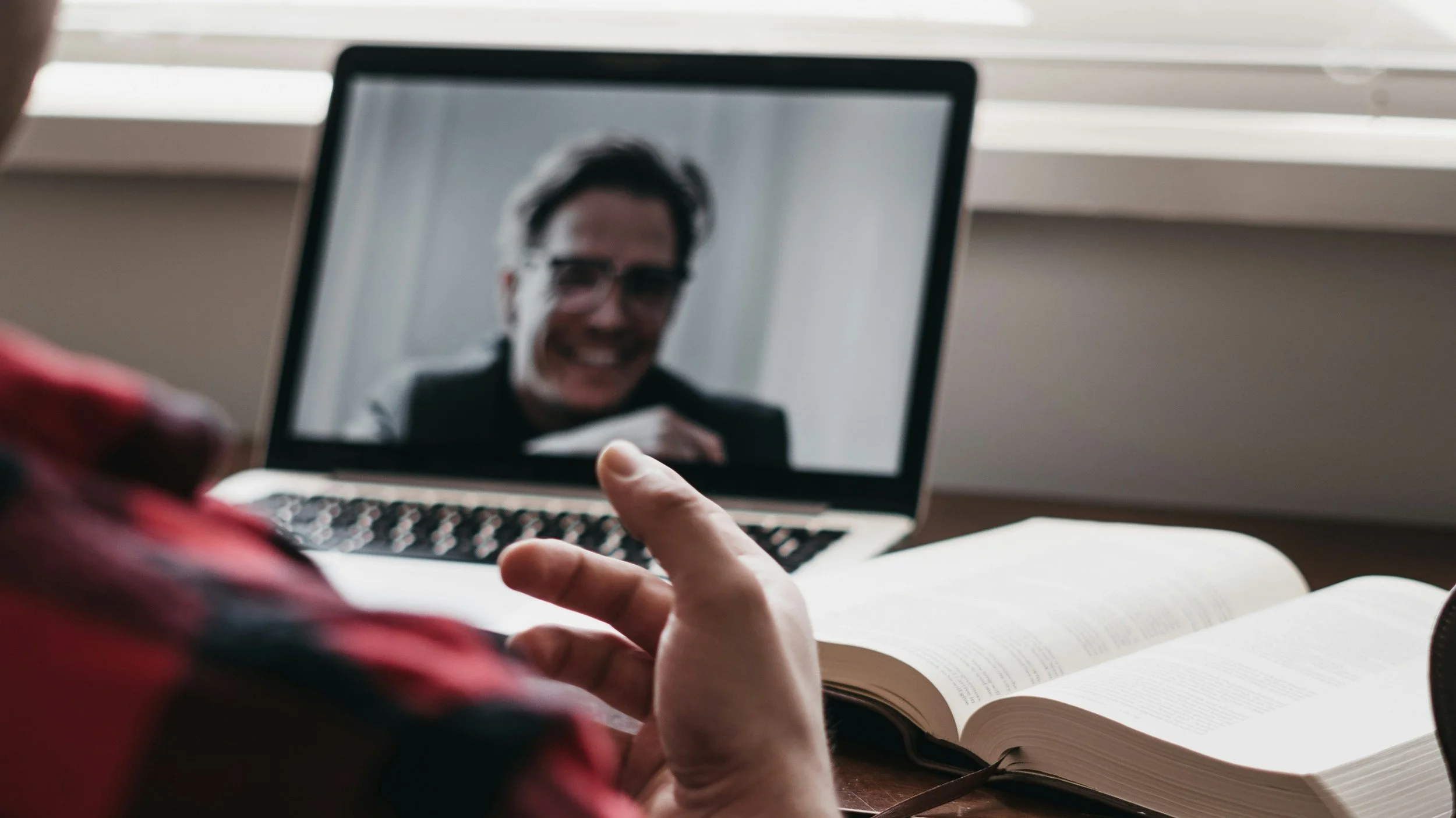 Person taking notes while on a video call on a laptop, with a book open on the table in front of them.
