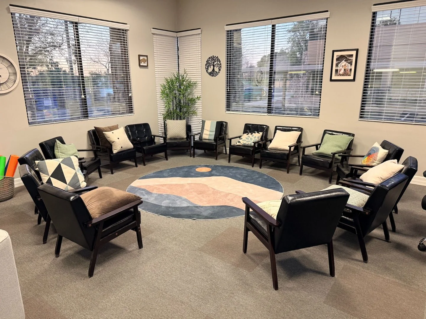 A circular arrangement of black chairs with various cushions in a well-lit room with large windows, a colorful round rug in the center, and wall decorations including framed pictures and a large decorative clock.