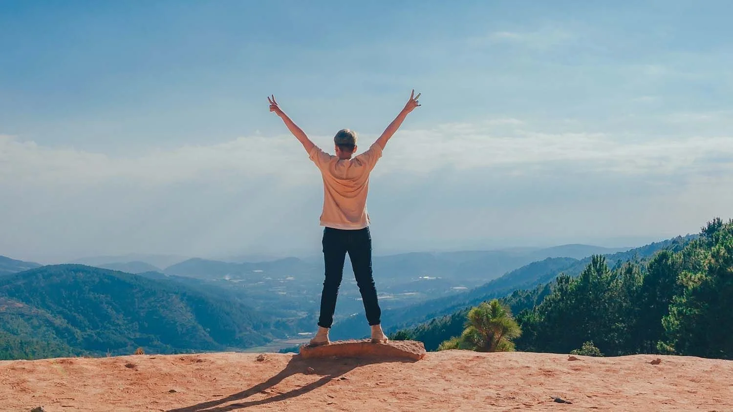 Person standing on a rock at a mountain overlook with arms raised, facing a scenic view of mountains and sky.